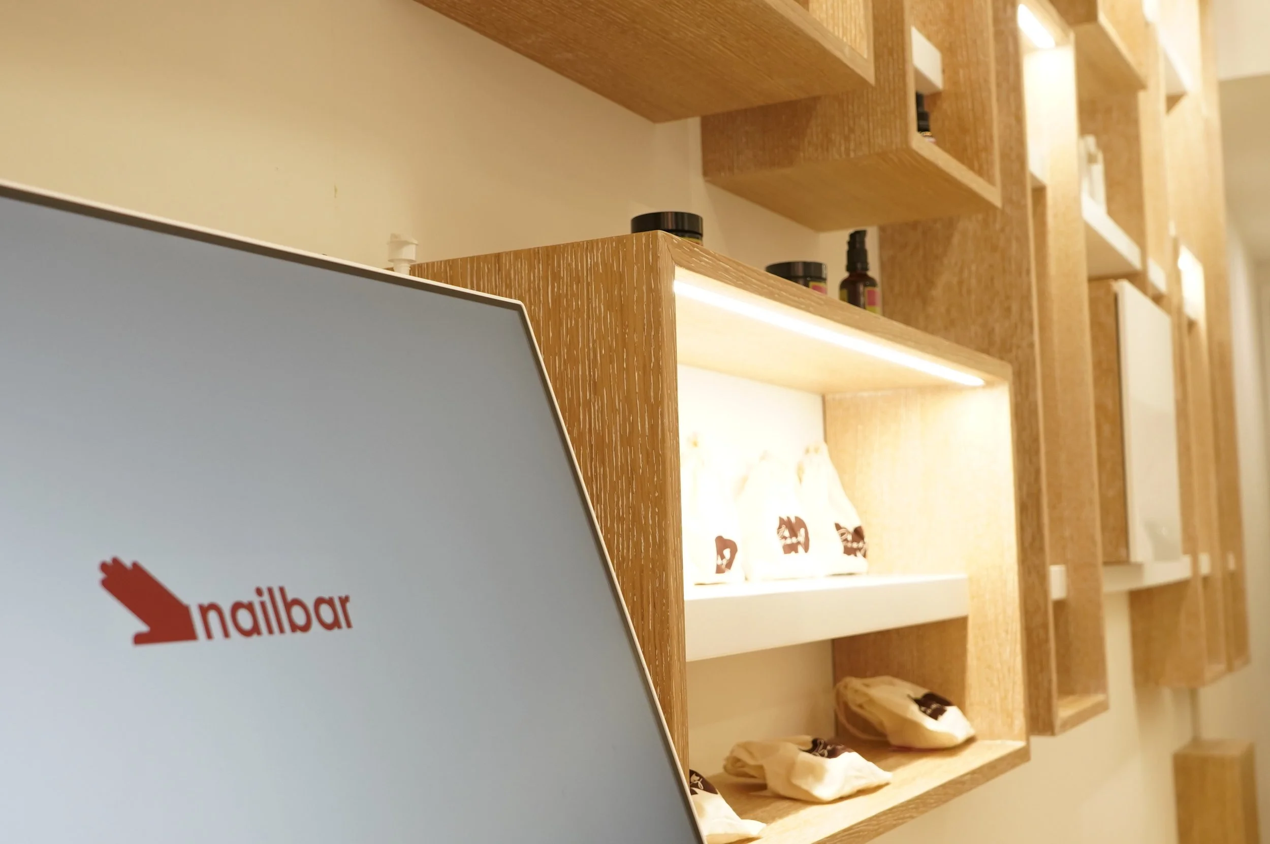 Particolare di uno scaffale di legno con vari oggetti e prodotti, e in primo piano una parte di uno schermo con il logo Nailbar.