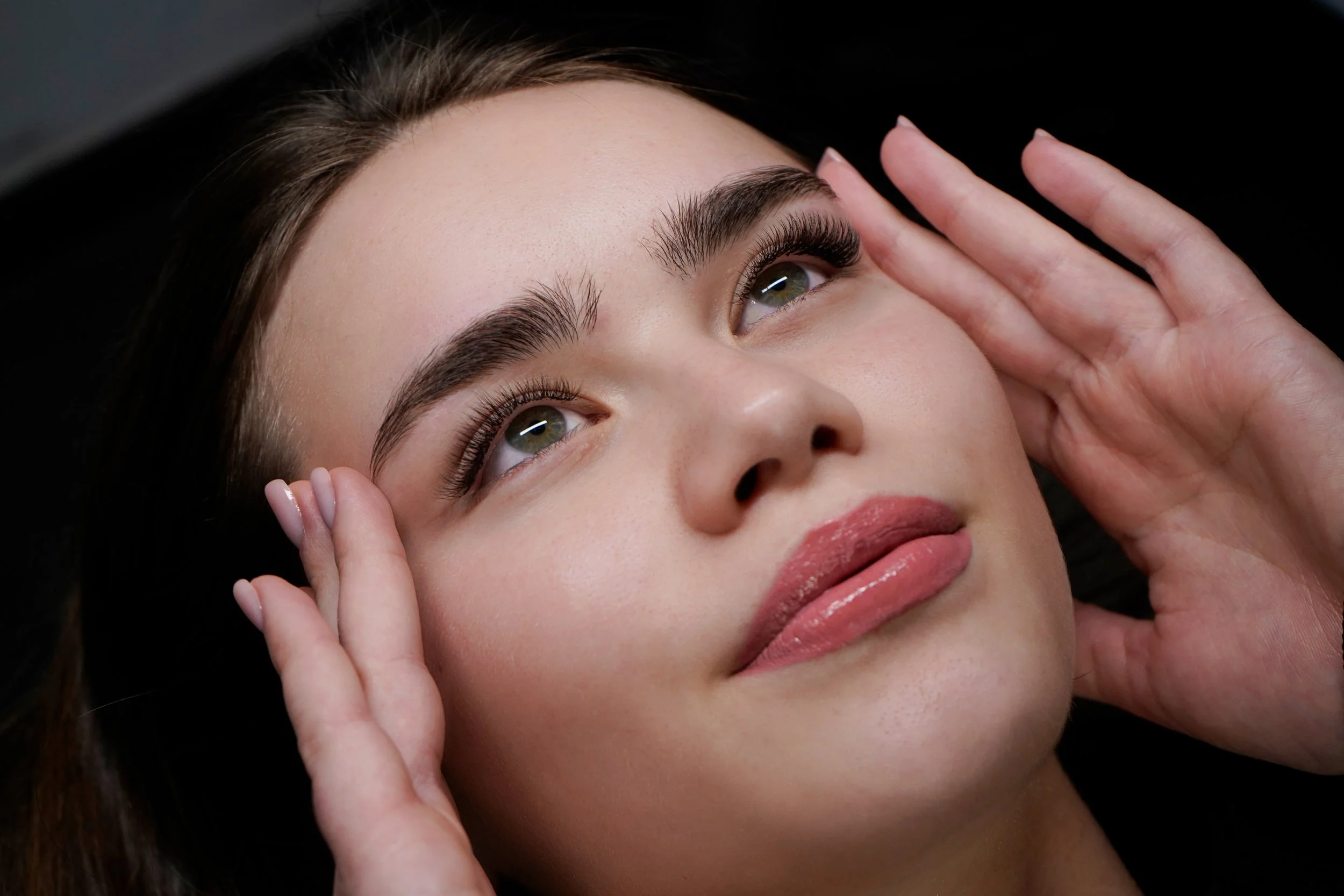 Primo piano di una donna con sopracciglia folte, occhi verdi e trucco leggero, mentre una mano si avvicina al suo viso.