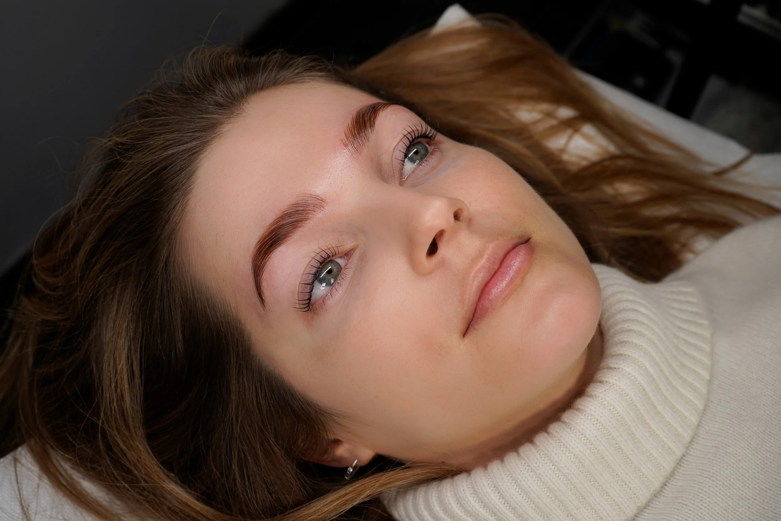 Ragazza con occhi azzurri e sopracciglia ben definite, che giace su una superficie, con capelli castani lunghi e ricci, indossa un maglione color crema.