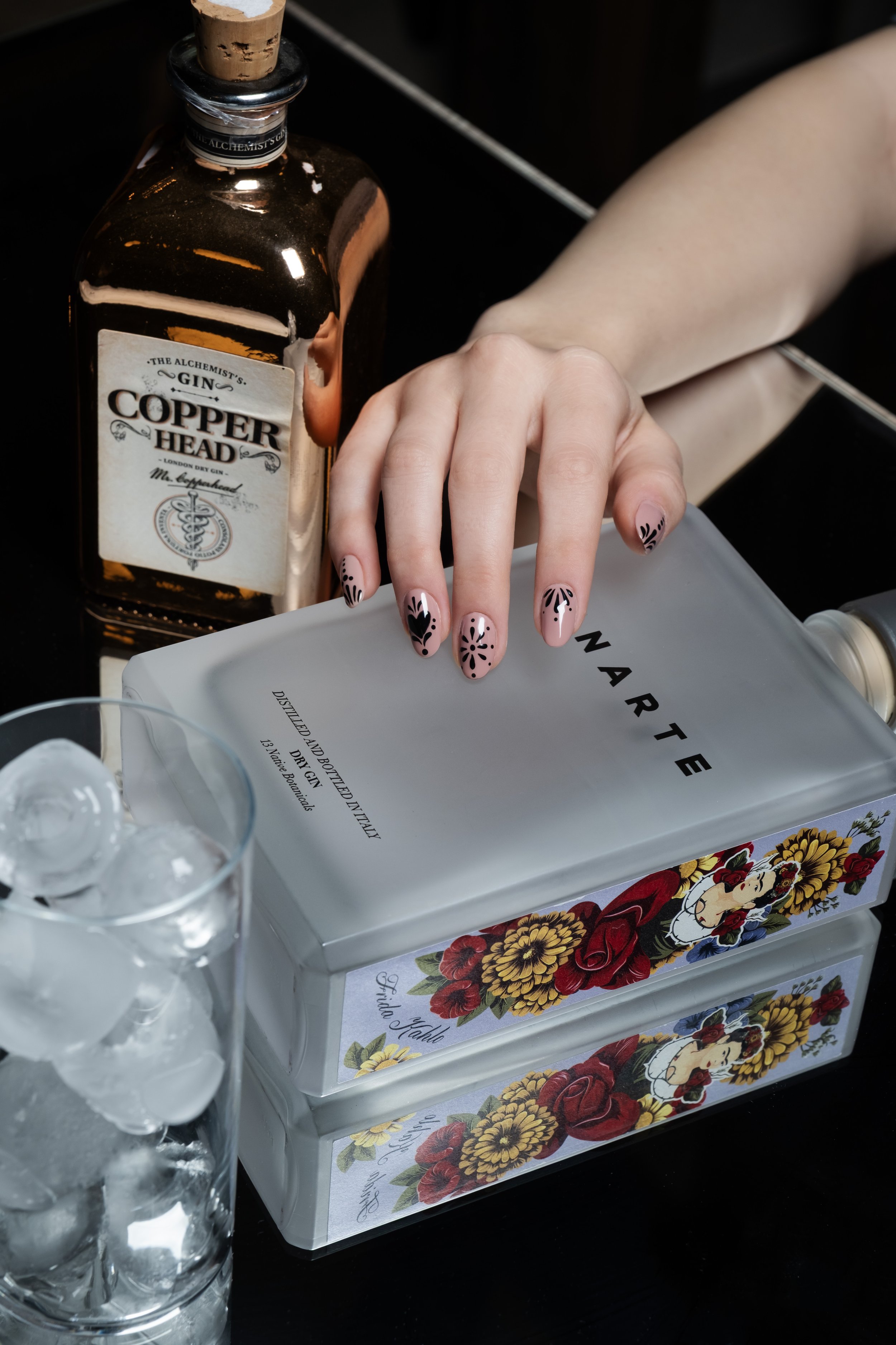 Una bottiglia di Gin Copper Head, un vaso di cristallo con cubetti di ghiaccio, e una scatola decorata con fiori e un'immagine di Frida Kahlo su un tavolo nero.