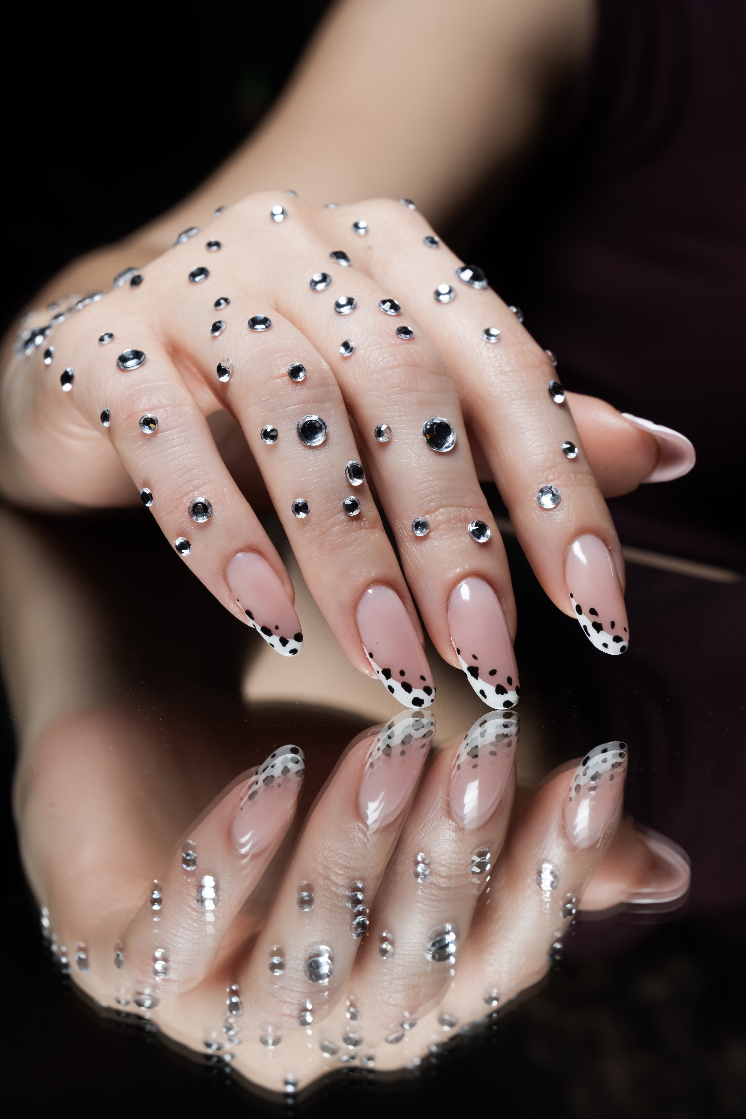 Unghie decorate con smalto trasparente e dots neri, con alcuni strass brillanti, riflesso delle mani su una superficie lucida.