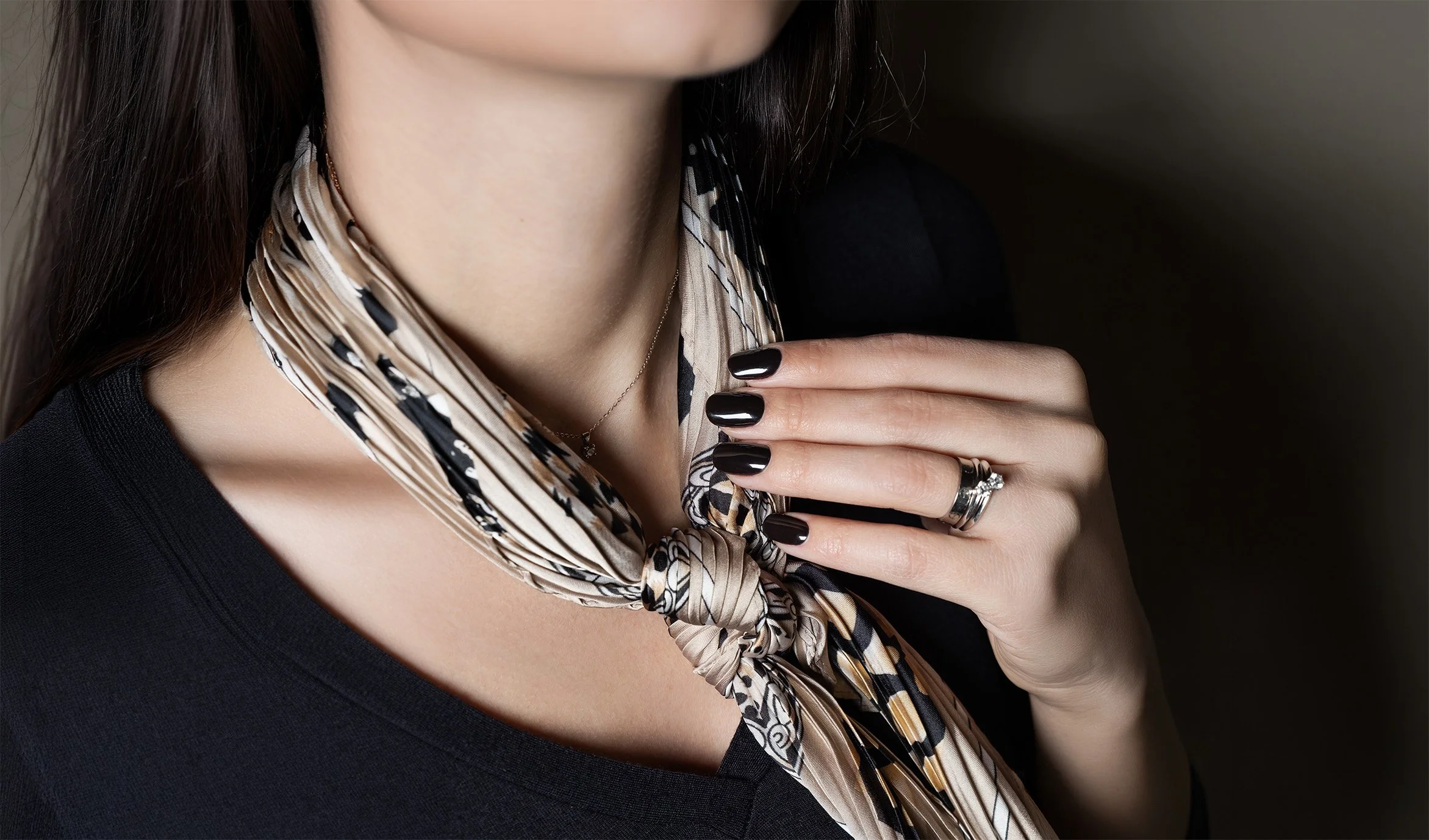 Una donna con un sottile collier d'oro, indossa un maglione nero, ha i capelli castani lunghi e lisci, e porta un foulard con motivo animalier annodato attorno al collo. La sua mano con un anello robusto e smalto nero è visibile, poggiata sul petto.