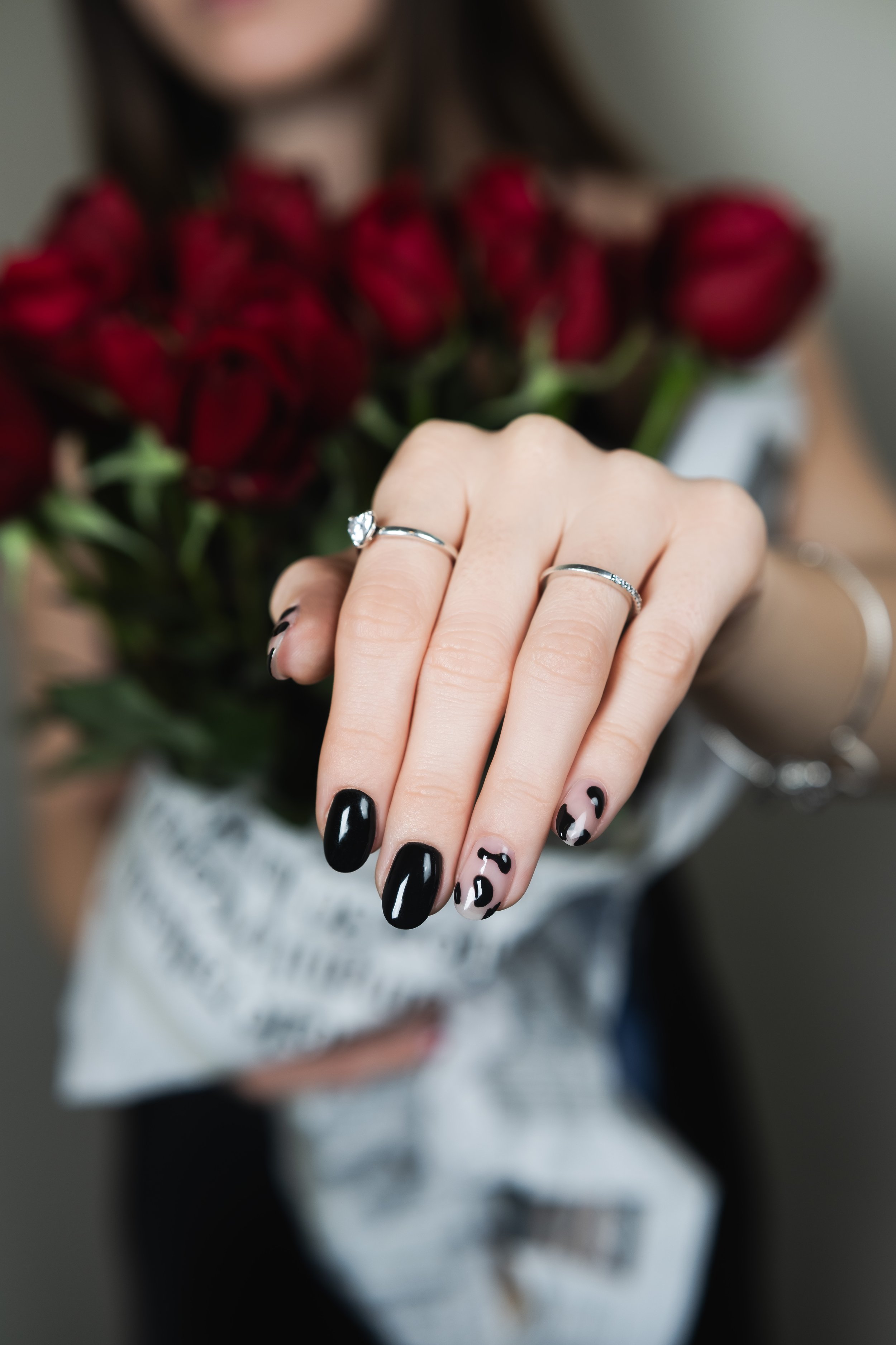 Mano con unghie nere e disegnate, che indossa due anelli, con bouquet di rose rosse sullo sfondo.