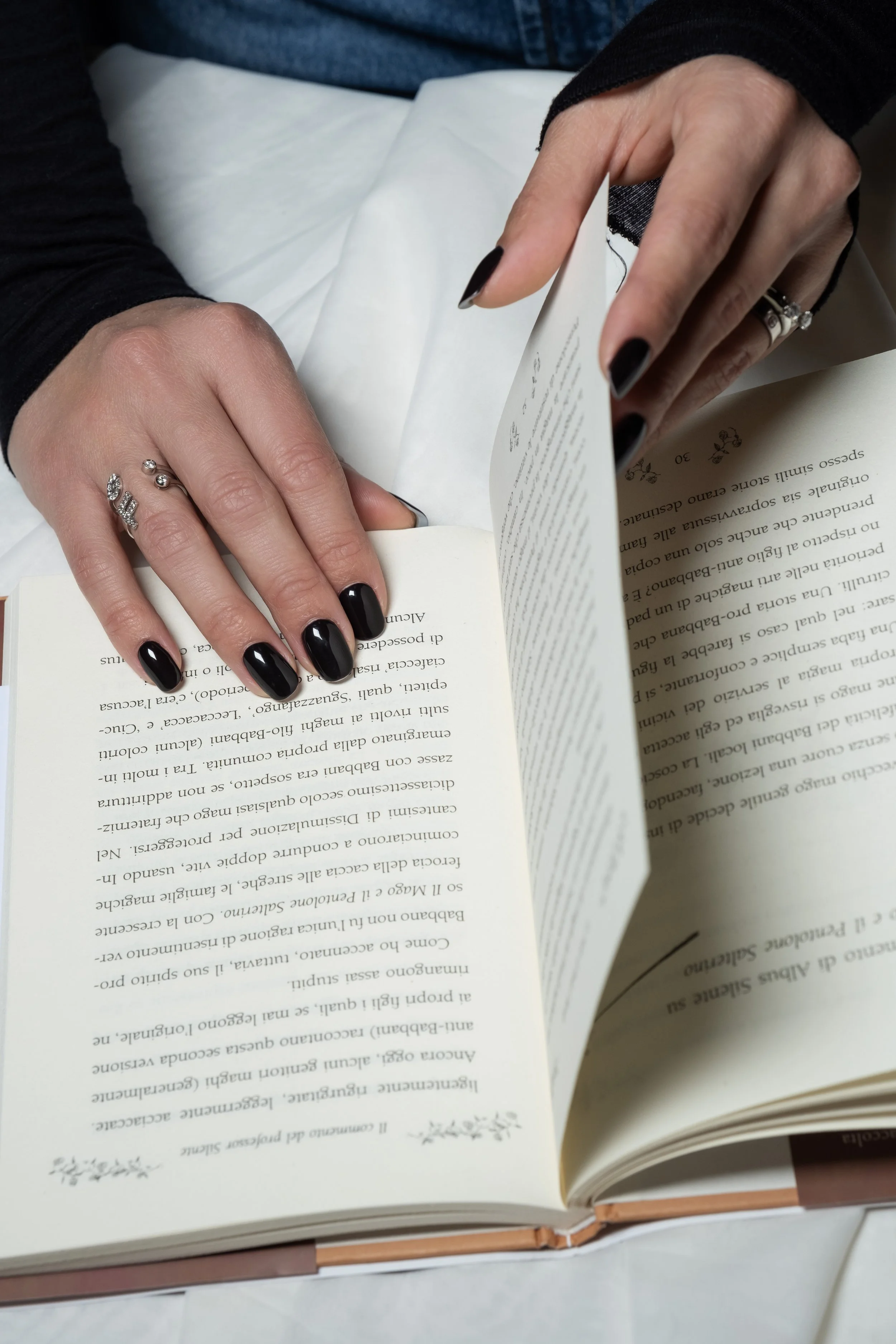 Una persona con le unghie dipinte di nero sta sfogliando un libro aperto, indossa anelli tra cui uno con pietra e un altro con scritte. Nelle vicinanze ci sono mani che tengono il libro.