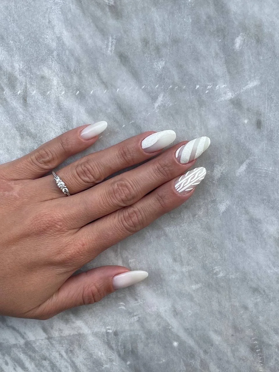 Unghie dipinte di bianco con motivi ondulati e linee sottili di colore argento, mano con anello con diamanti, su sfondo di superficie grigia.