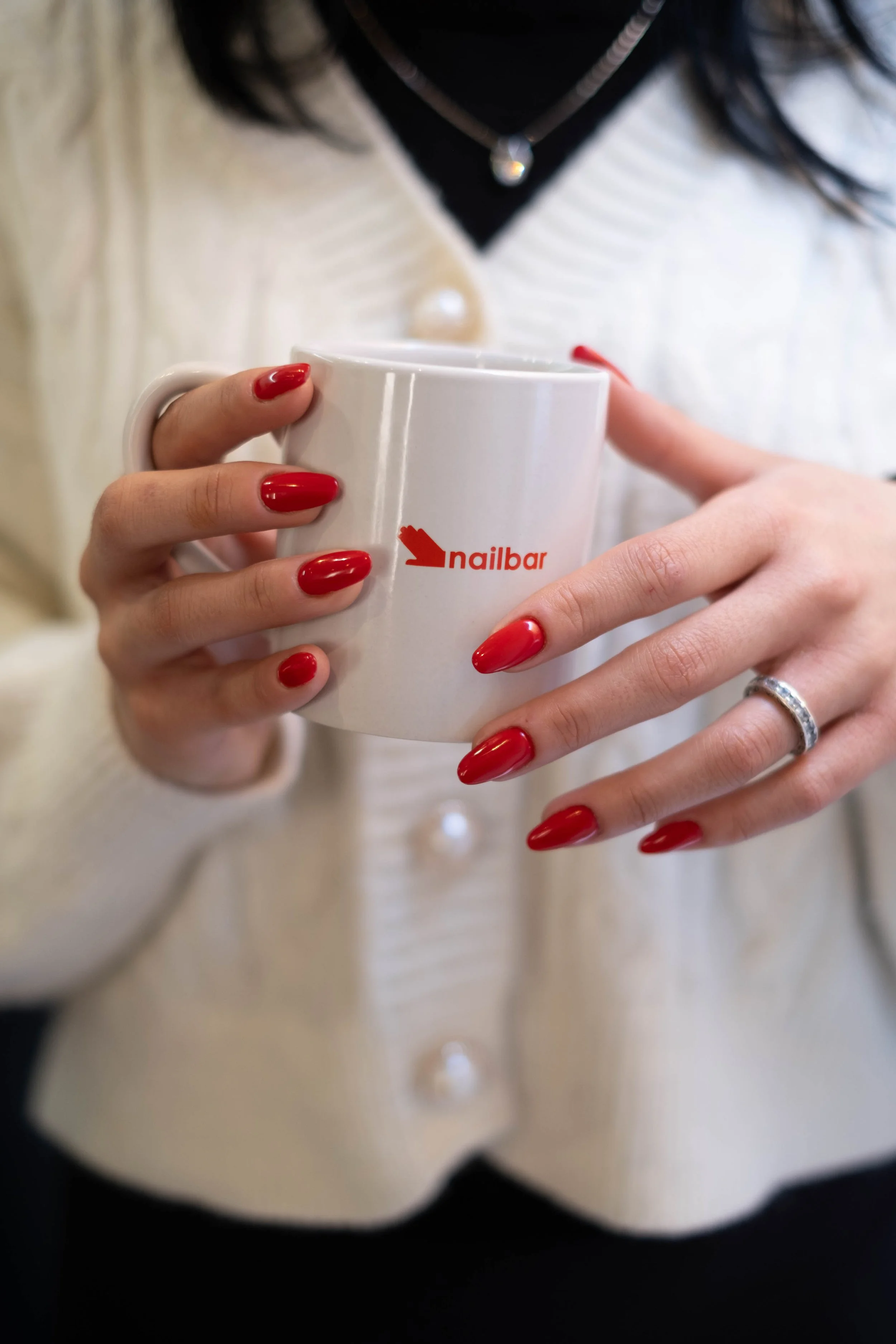 Una persona tiene le mani con un smalto rosso brillante che tiene una tazza bianca con il logo Nailbar. La persona indossa un anello e ha anche un monile al collo.