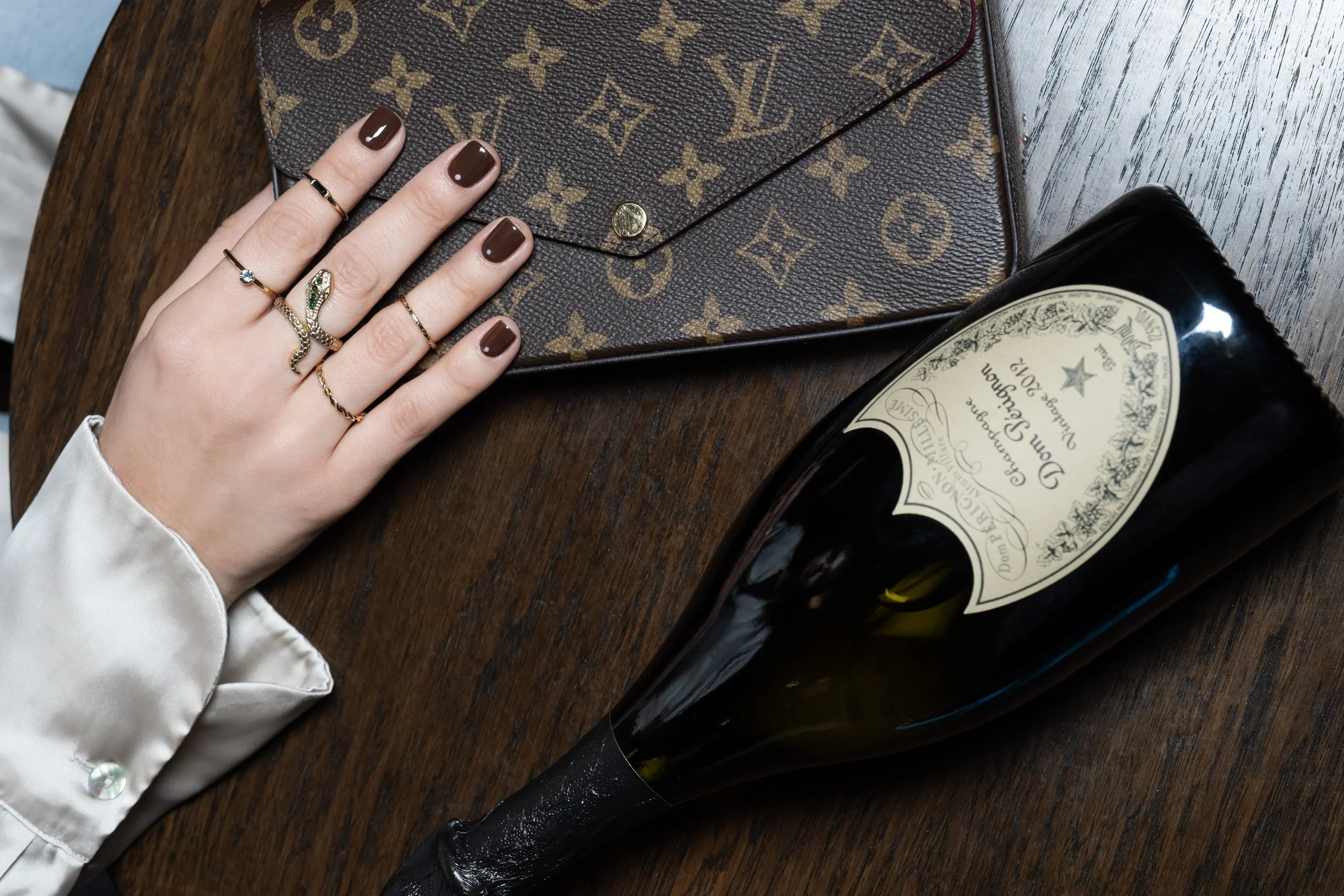 Un portafoglio marrone con motivo Louis Vuitton, alcune mani con anelli e smalto marrone scuro, una bottiglia di champagne con etichetta vintage, tutti posti su un tavolo di legno.