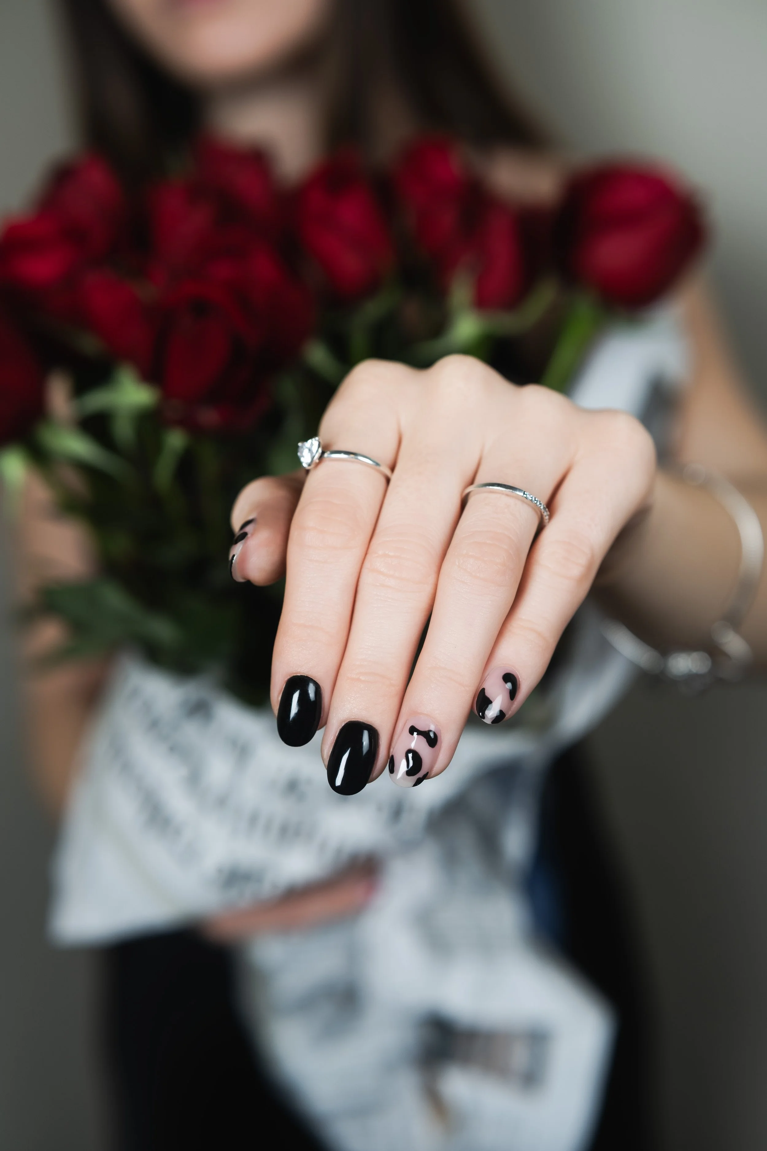 Mano con un anello di diamanti e unghie decorate con smalto nero e disegni leopardati, con un mazzo di rose rosse sullo sfondo.
