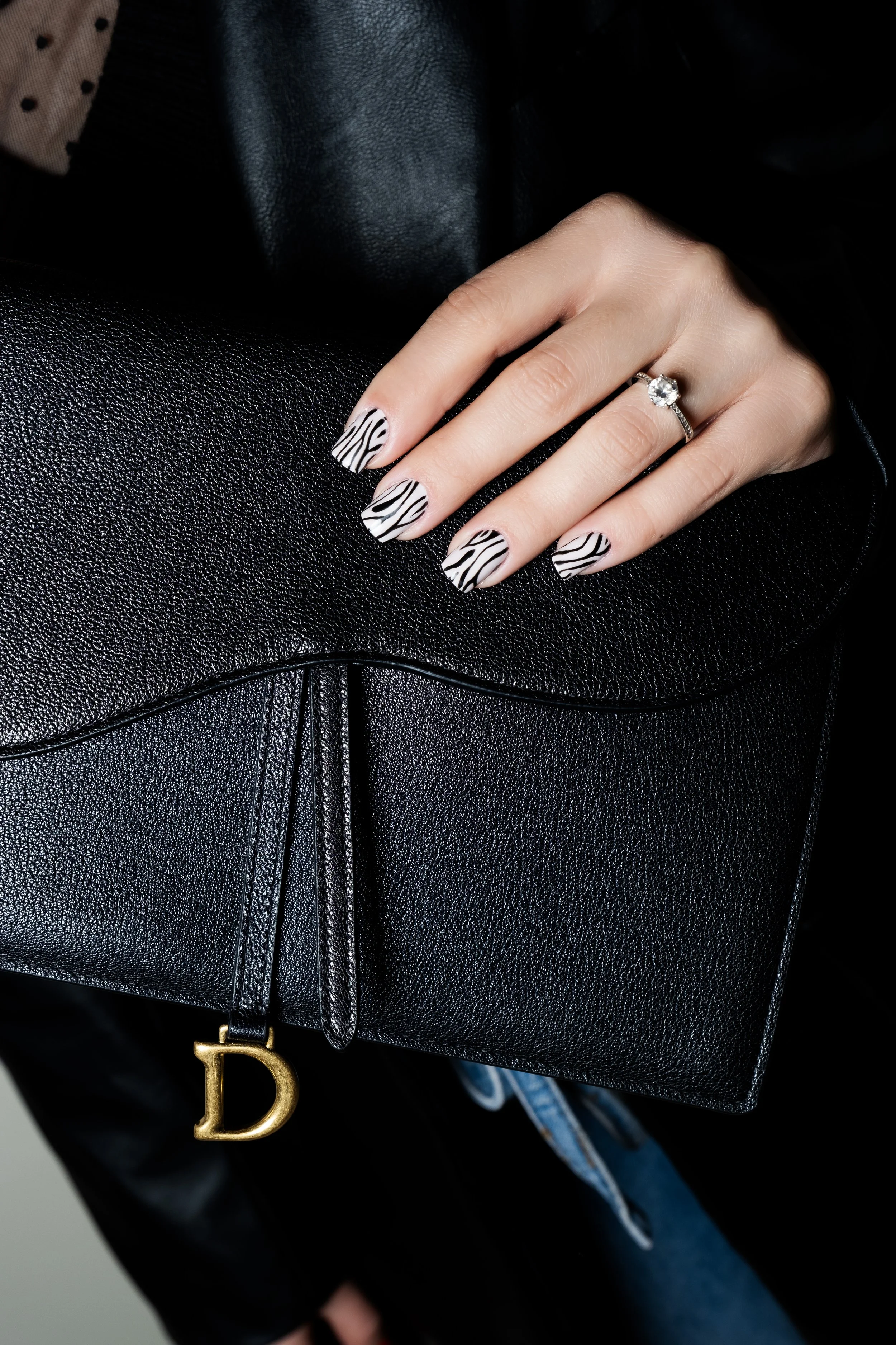 Una mano con unghie dipinte con motivo zebra, indossando un anello con pietra brillante, appoggiata su una borsa nera in pelle con la penna attaccata alla cerniera.