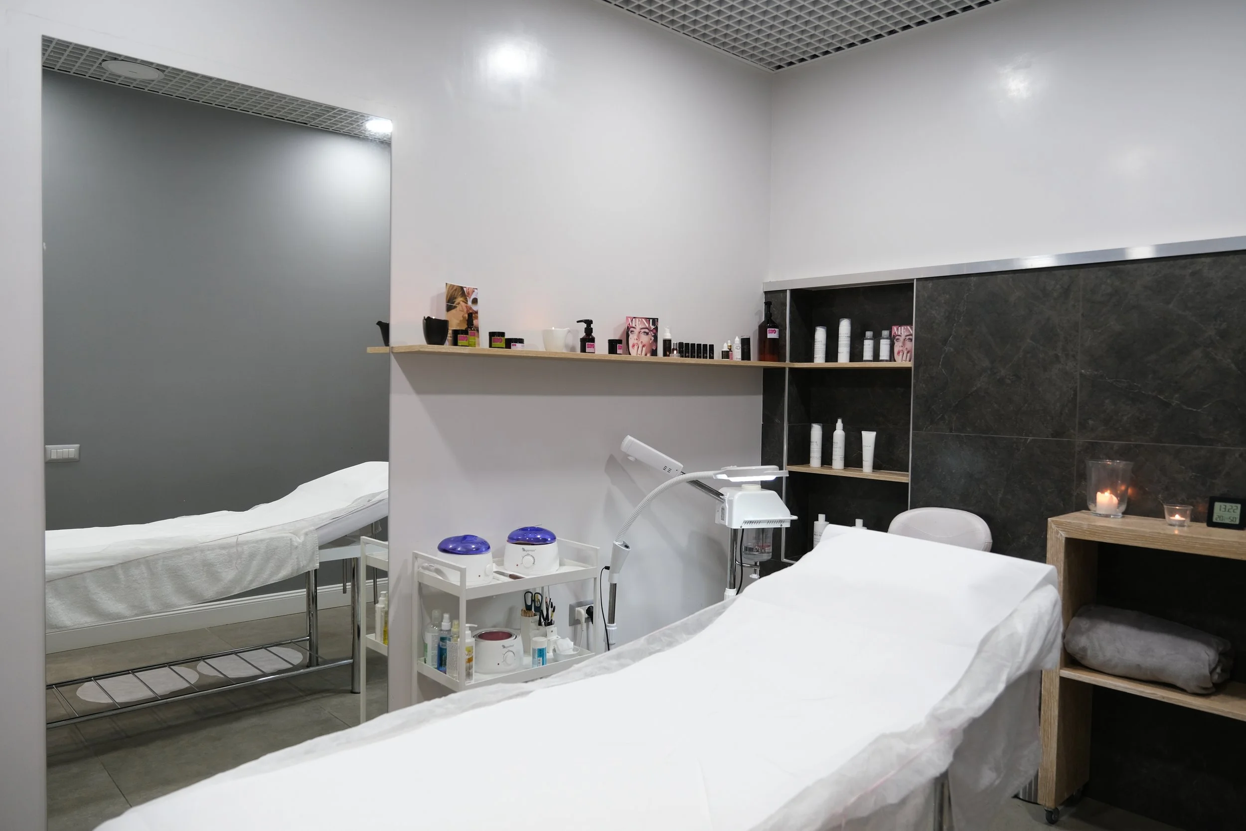 Studio estetico con lettini, scaffale con prodotti di bellezza, strumenti per trattamenti estetici, e decorazioni come candele e poster, ambiente moderno e minimalista.