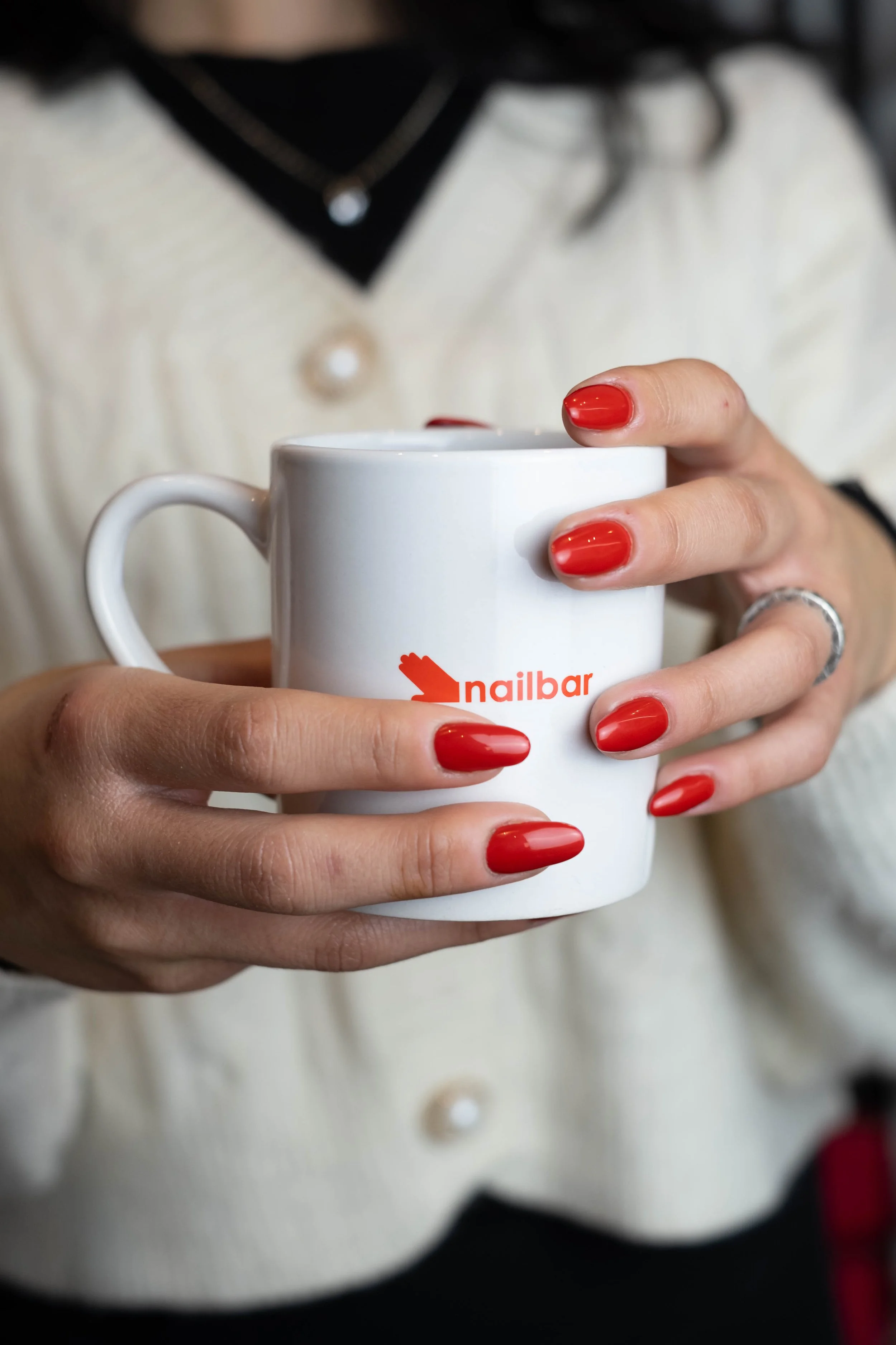 Una persona che tiene una tazza bianca con il logo 'nailbar' e un fiore rosso, con unghie rosse lucide, indossa un maglione bianco con decorazioni di bottoni e un reggiseno nero con dettagli di perle e collane.