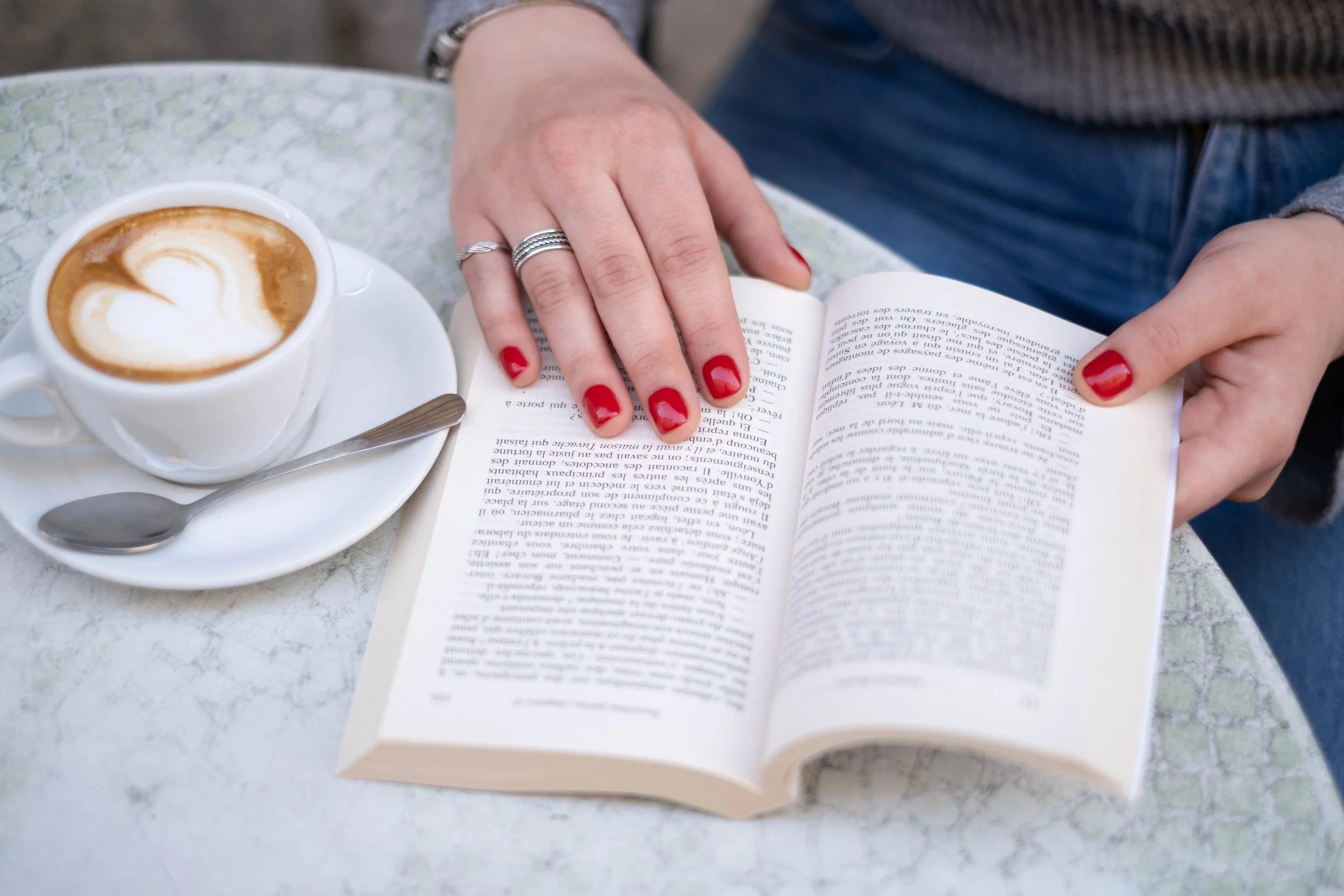 Una tazza di caffè con latte decorato con cuore, una mano con unghie rosse reading un libro aperto seduti a un tavolo di marmo.