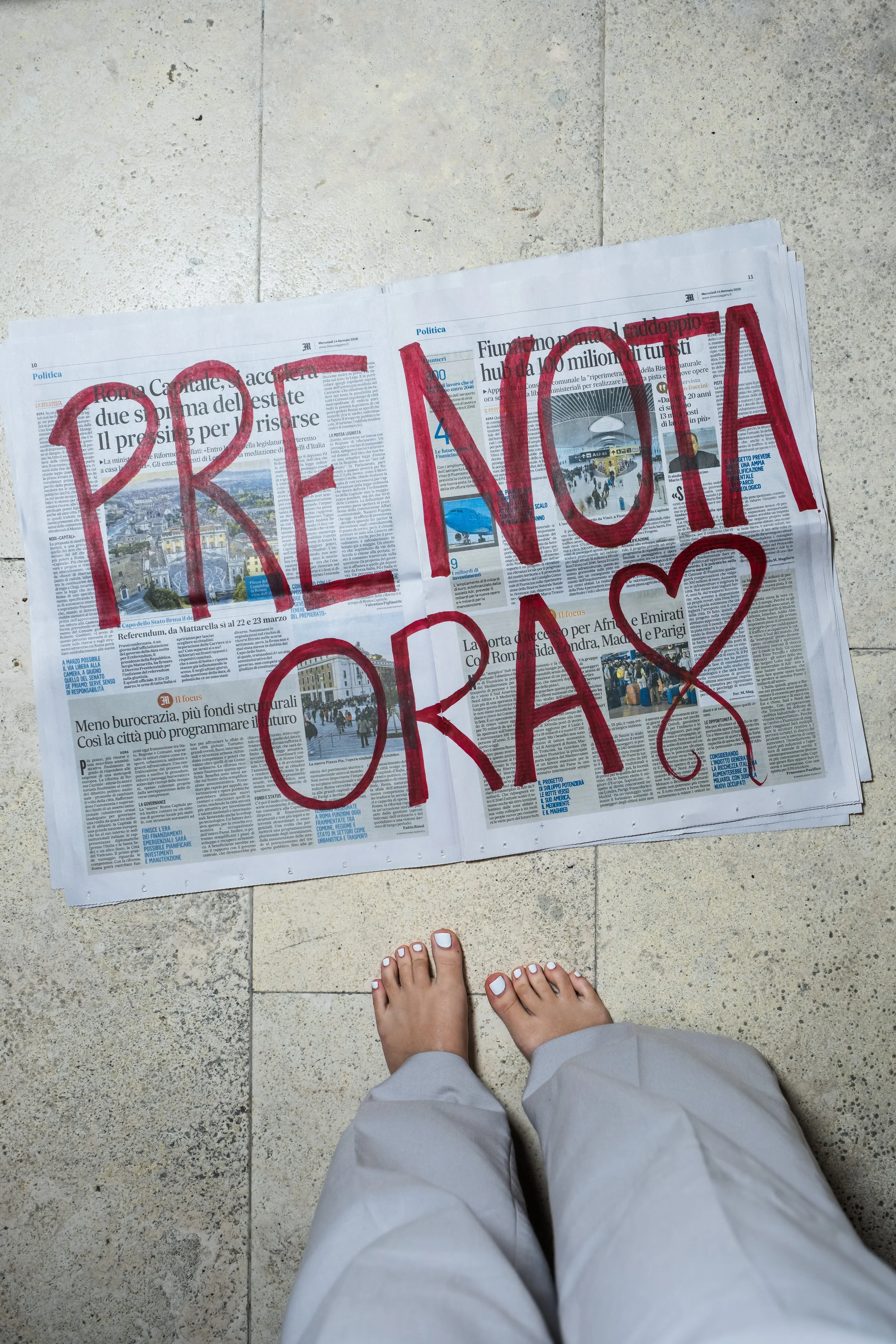 Un giornale appoggiato sul pavimento con scritto a mano 'FRENO OME' in rosso con un cuore disegnato. In primo piano ci sono i piedi di una persona con le unghie dipinte di bianco e pantaloni di colore chiaro.
