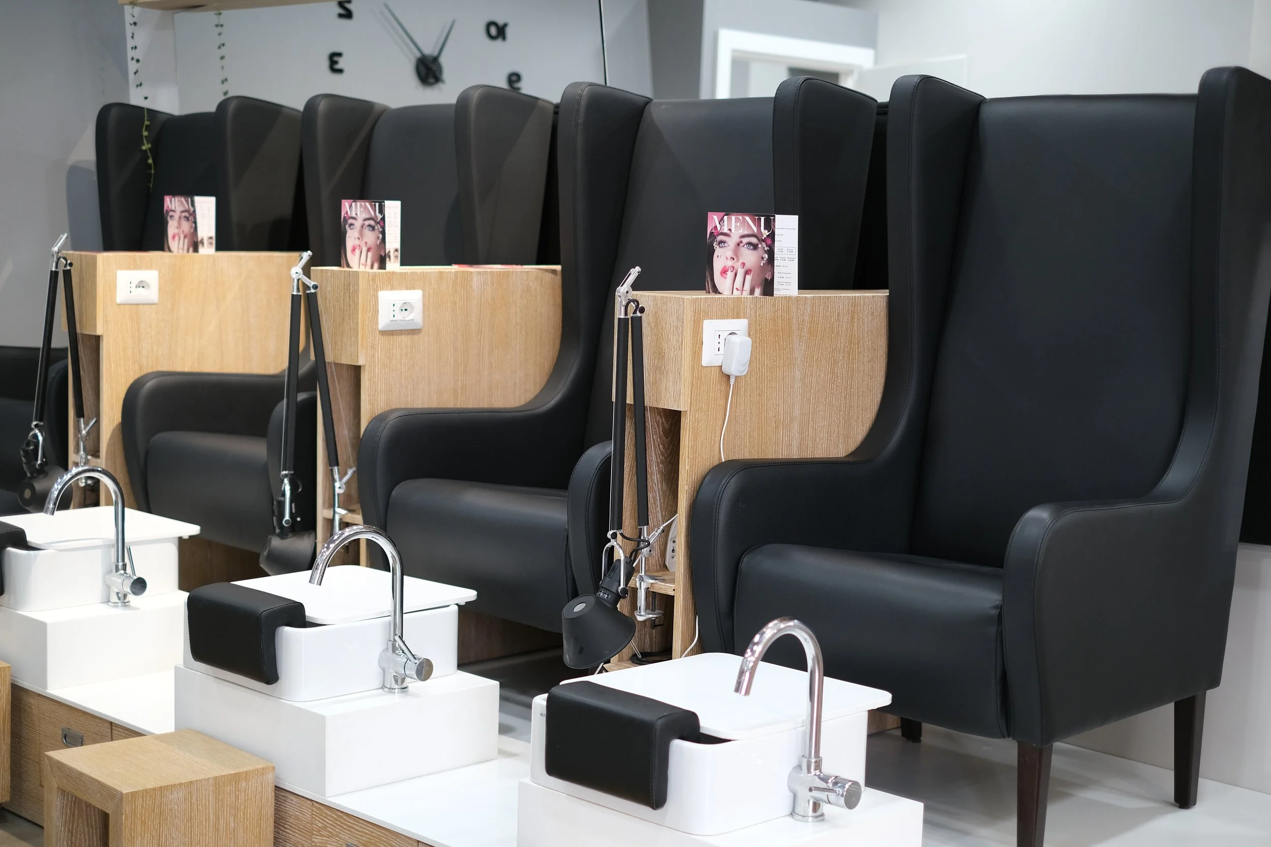 Stazioni di lavaggio per i piedi con sedili neri e divanetti in legno, con menu visibili e prese di corrente, in un salone di un centro di bellezza o salone di parrucchiere.