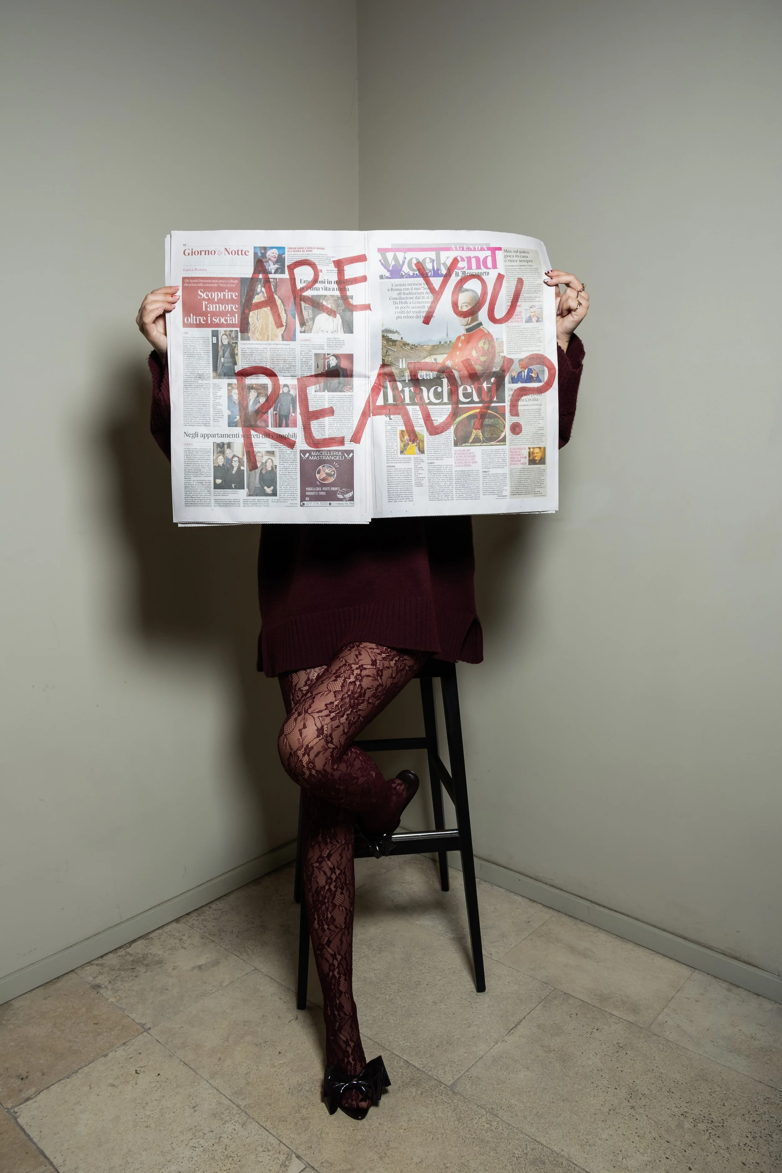 Persona vestita con calze a rete rosse, gonna e stivali alti neri, che si appoggia a una sedia e tiene un giornale davanti al volto con scritto 'Are you crazy?' in rosso