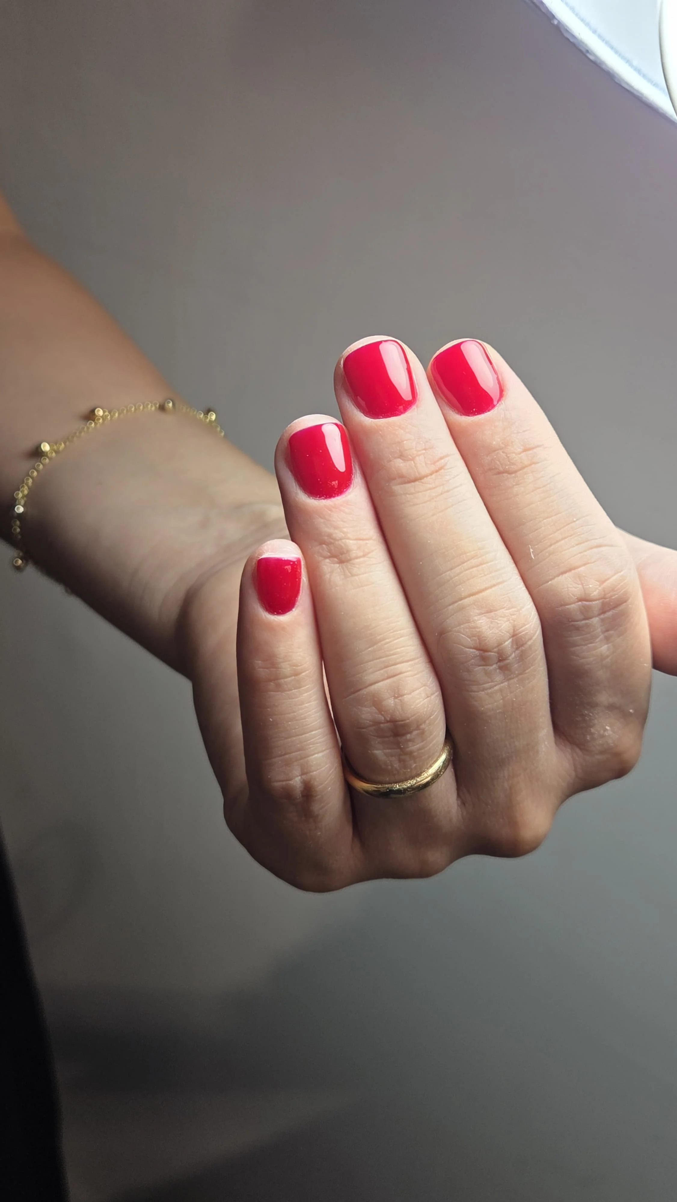 Una mano con unghie dipinte di rosso lucido, indossa due anelli dorati, uno sulla mano e uno sul dito anulare, con un bracciale sottile dorato.
