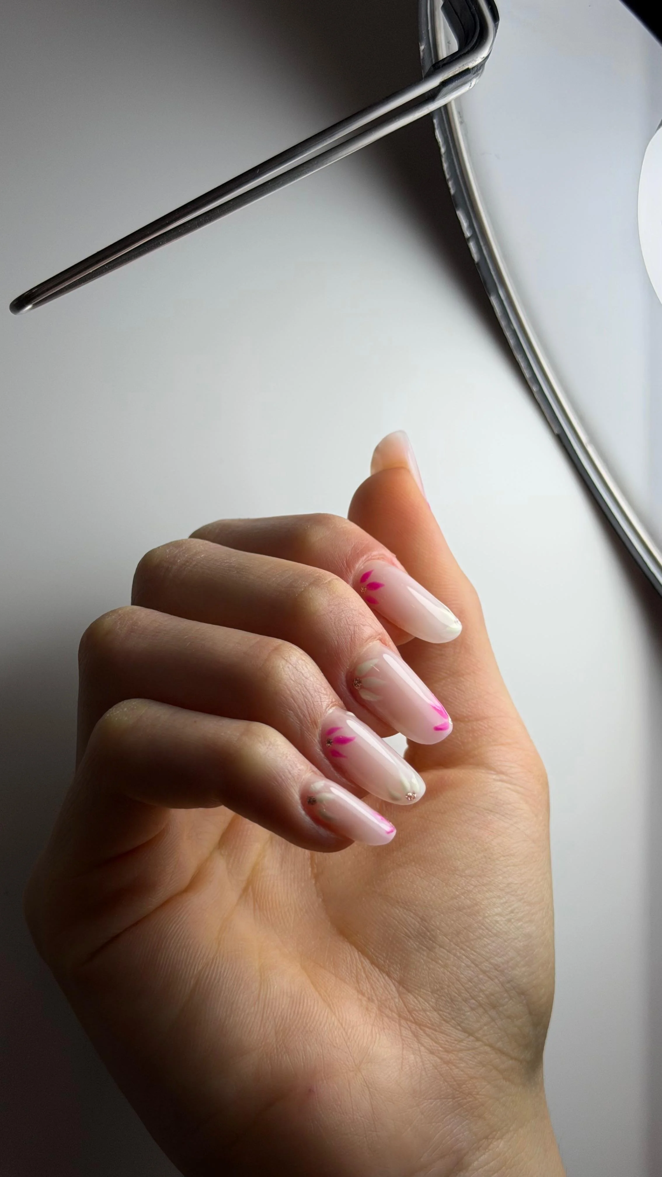 Mano con unghie decorate con smalto rosa e dettagli dorati, riposta su tavolo bianco accanto a uno specchio