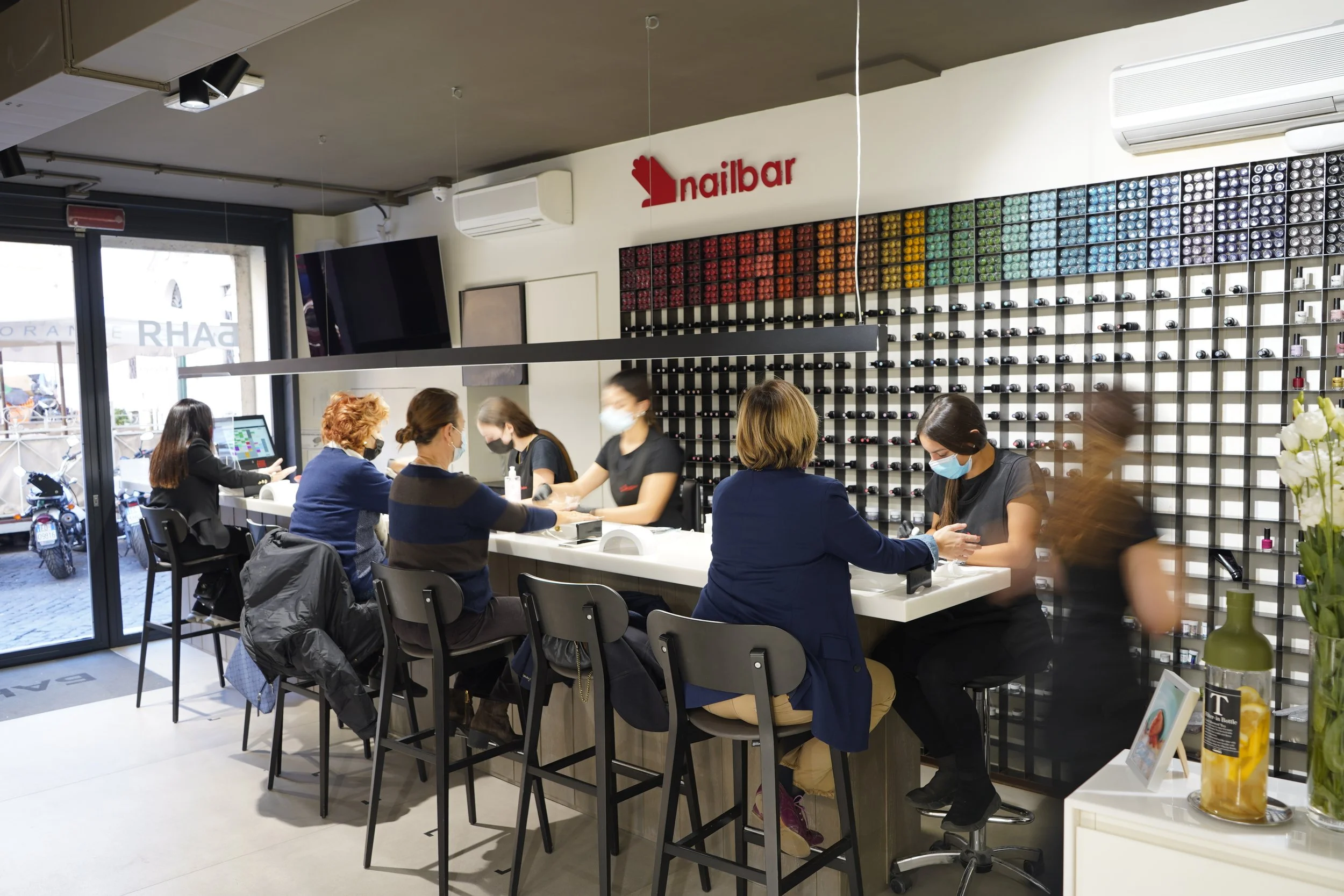 Interno di un salone di bellezza con clienti e estetisti, parete con scaffale di smalti per unghie, logo 'nailbar' sulla parete e grandi finestre che permettono l'ingresso di luce naturale.