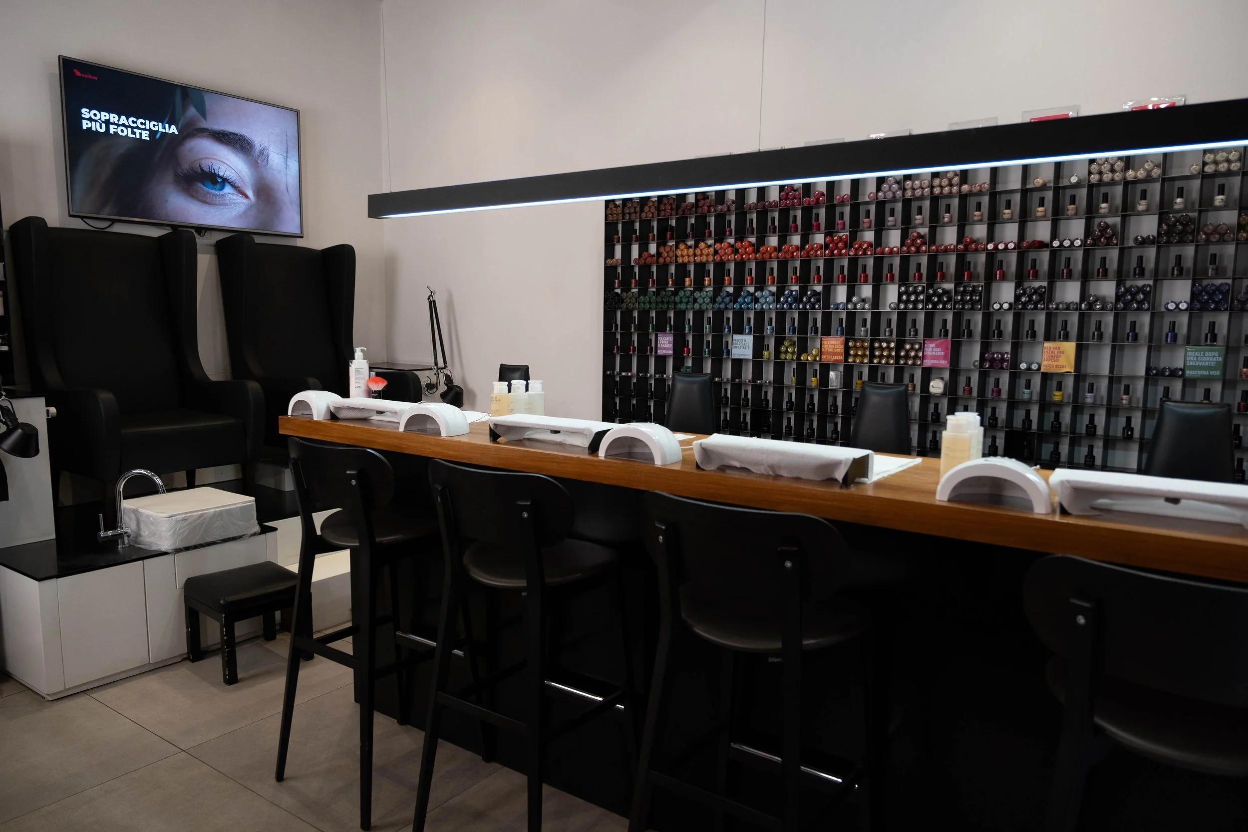 Interno di un salone di bellezza con parete di smalti per unghie e poltrone da manicure, tavolo con strumenti, schermo pubblicitario e scaffale con smalti colorati.
