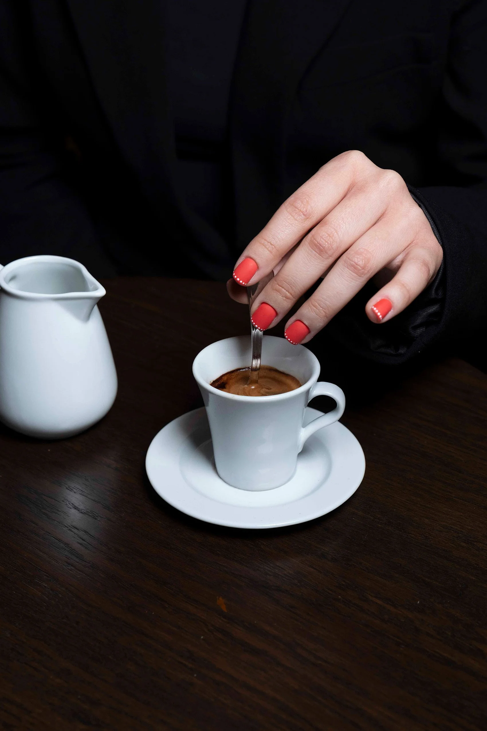 Mano con unghie laccate di rosso con decorazioni di puntini bianchi, che si immerge in una tazza di caffè su un tavolo di legno scuro, con una brocca di latte bianca sullo sfondo.