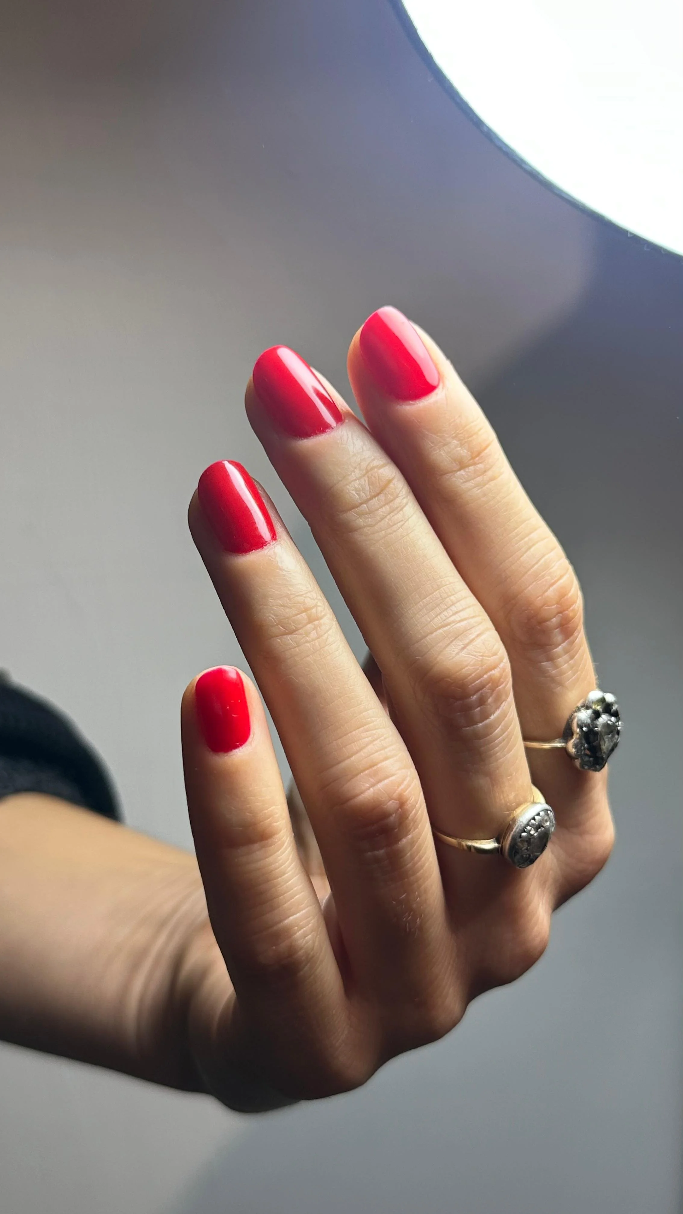 Dita di una mano con unghie dipinte di rosso, indossa due anelli con pietre, sfondo sfocato con una luce brillante in alto a destra.