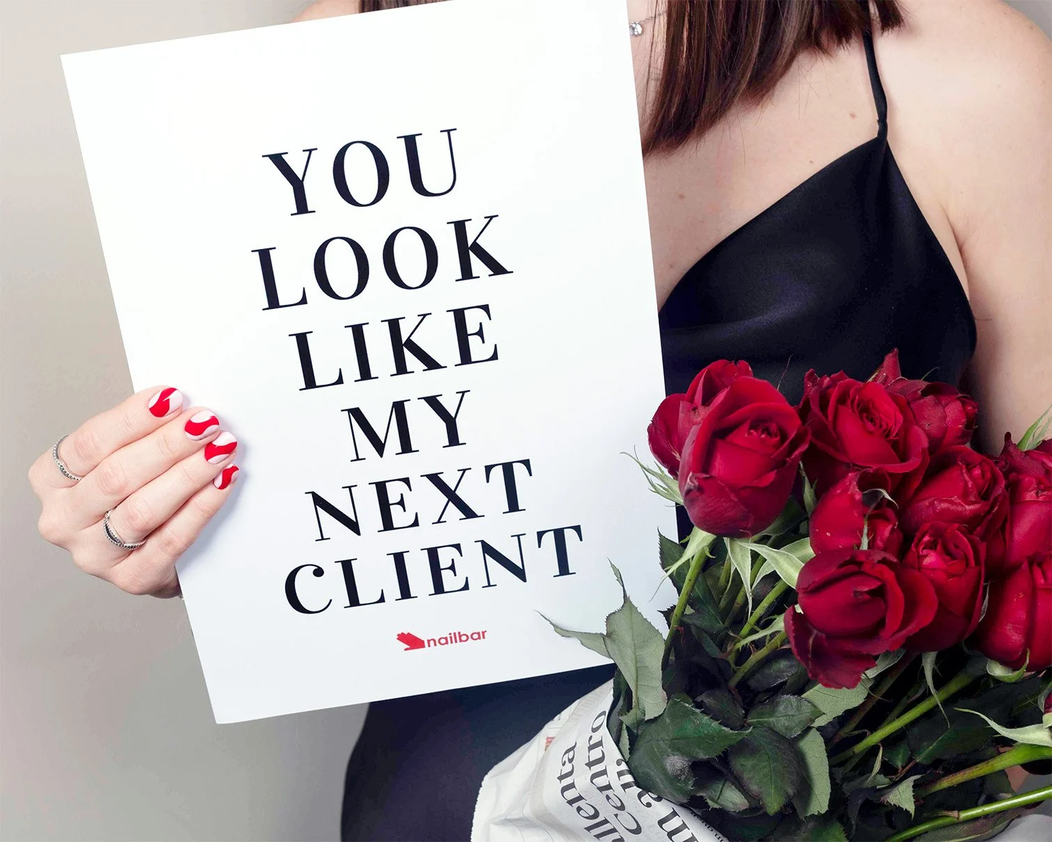 Una donna tiene un cartello con la scritta 'You look like my next client'. Accanto a lei c'è un mazzo di rose rosse e le sue mani hanno unghie con smalto rosso e bianco e indossa anelli argentati.