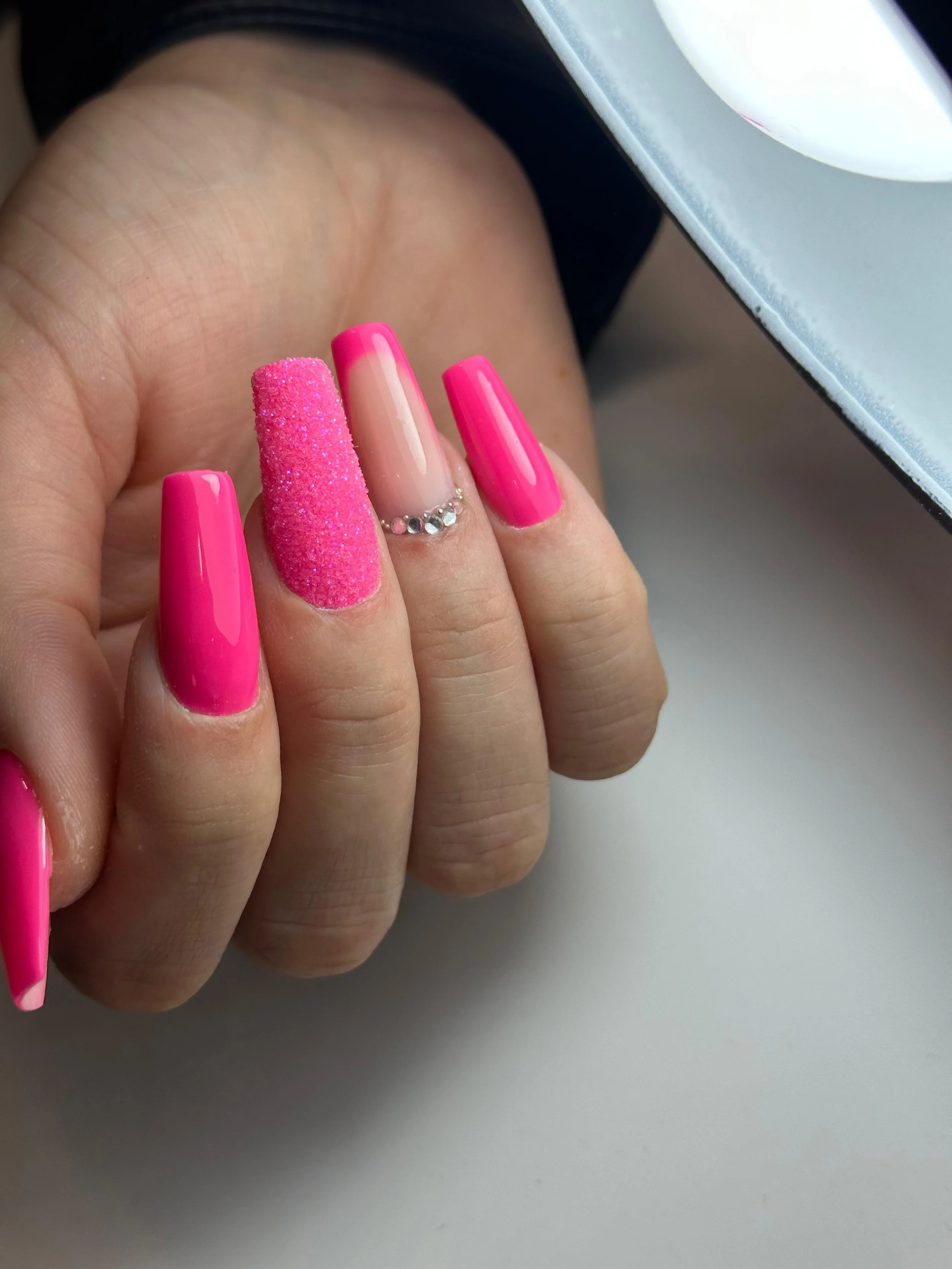 Unghie decorative con smalto rosa brillante e glitter, una con dettagli di cristalli, mano su tavolo con specchio sopra.