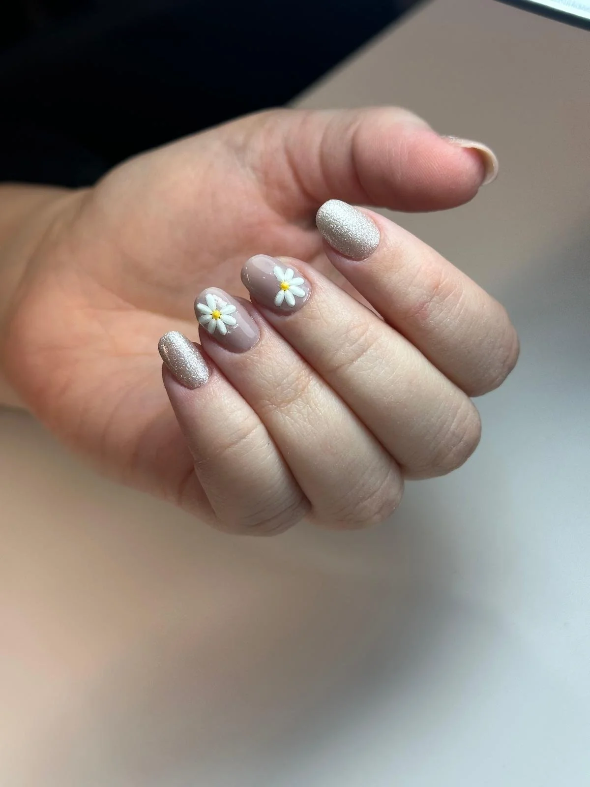 Un'unghia con decorazione floreale di margherite bianche e gialle, altre unghie color panna con glitter