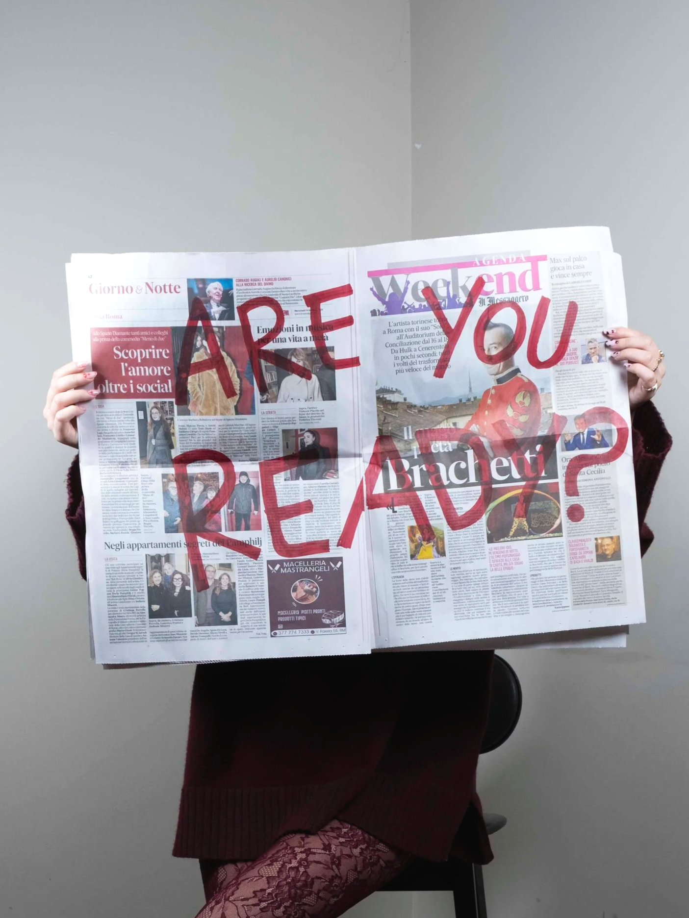 Una persona tiene in mano un giornale aperto con scritte rosse che dicono "ARE YOU READY?" sopra. La persona indossa un maglione marrone e le calze a rete rosse. Sul volto indossa una maschera nera. Lo sfondo è di colore grigio.