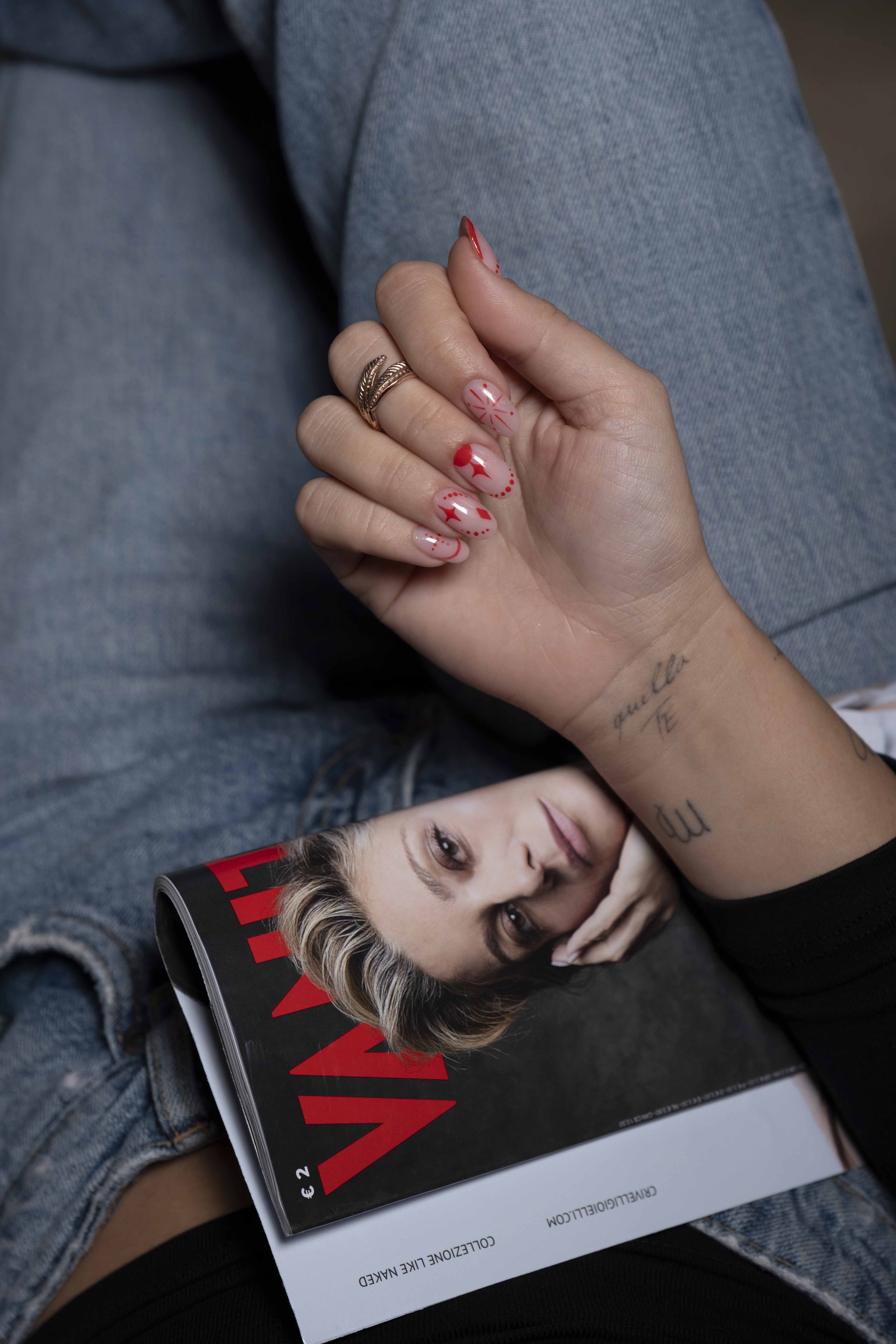 Una mano con unghie decorate con disegni rossi e glitter tiene in mano una rivista di moda con la copertina di una donna con capelli biondi e trucco elegante, appoggiata sulle gambe di una persona che indossa jeans.