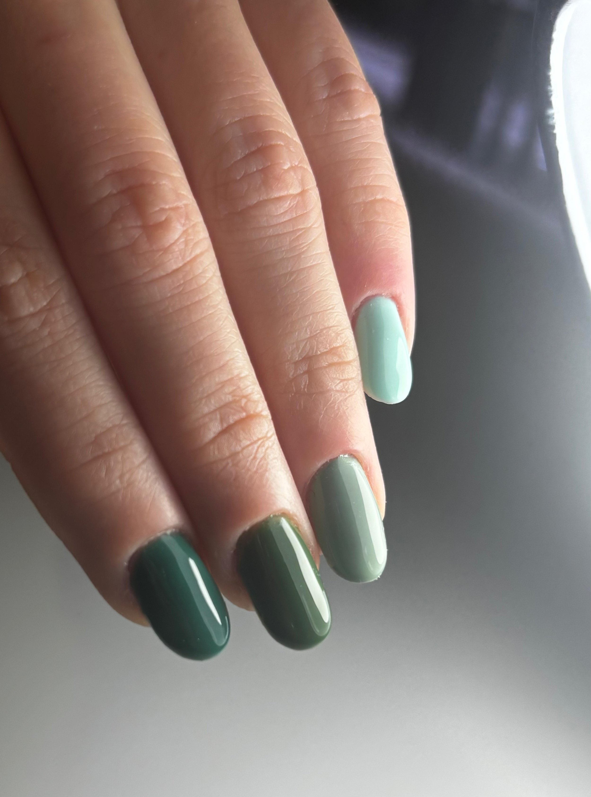 Dita di mani con unghie lunghe e curate, con smalto verde chiaro, sullo sfondo uno sfondo sfumato di grigi e neri.
