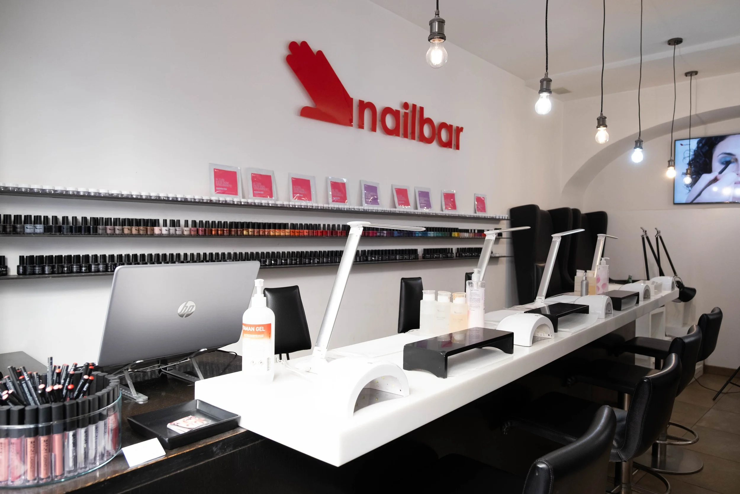 Interno di un negozio di nail art con vari espositori di smalti per unghie, postazioni per manicure, e illuminazione con lampadine a vista.