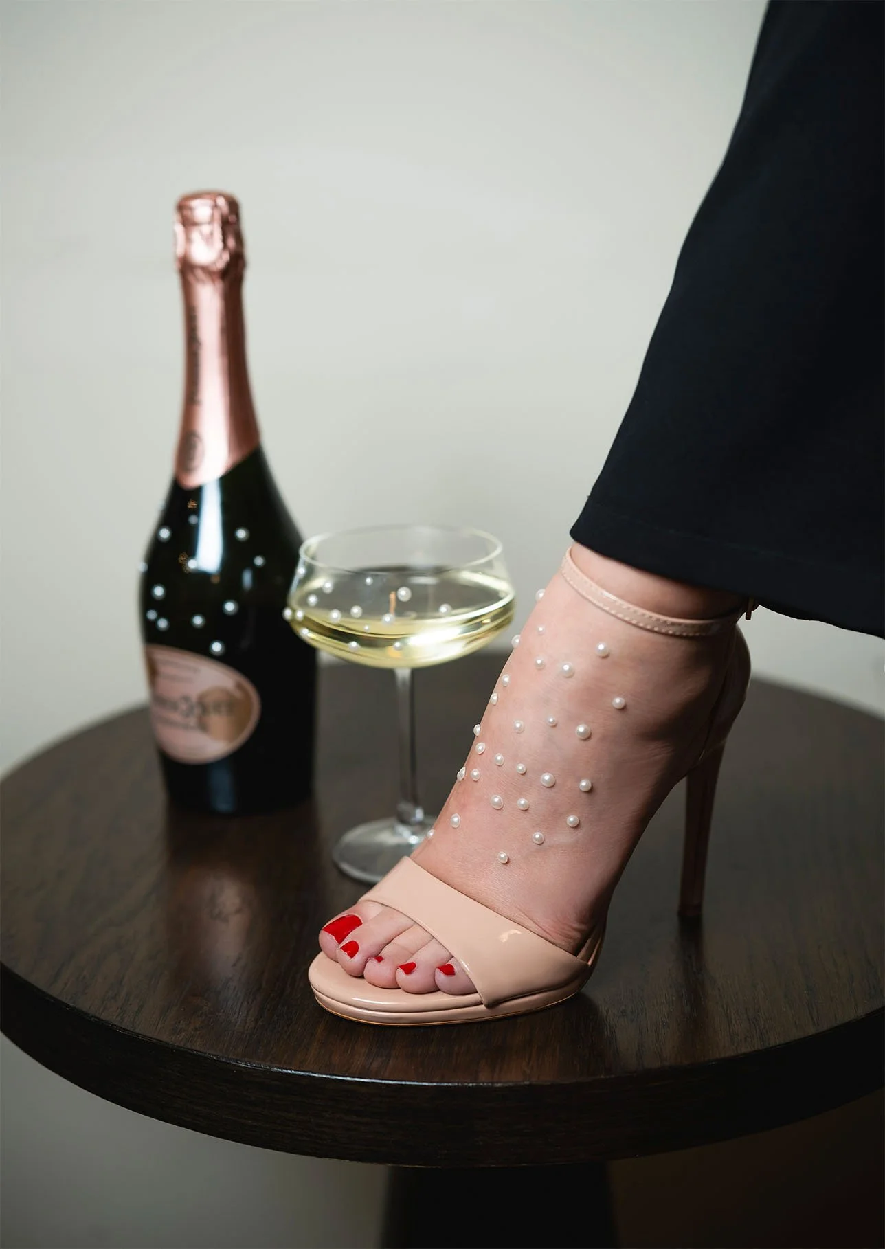 Una donna indossa tacchi a spillo beige decorati con perle, con le dita dei piedi dipinte di rosso. Sul tavolo di fronte c'è una bottiglia di champagne rosa, un bicchiere di champagne e una gamba coperta da una calza trasparente con dettagli di perle.