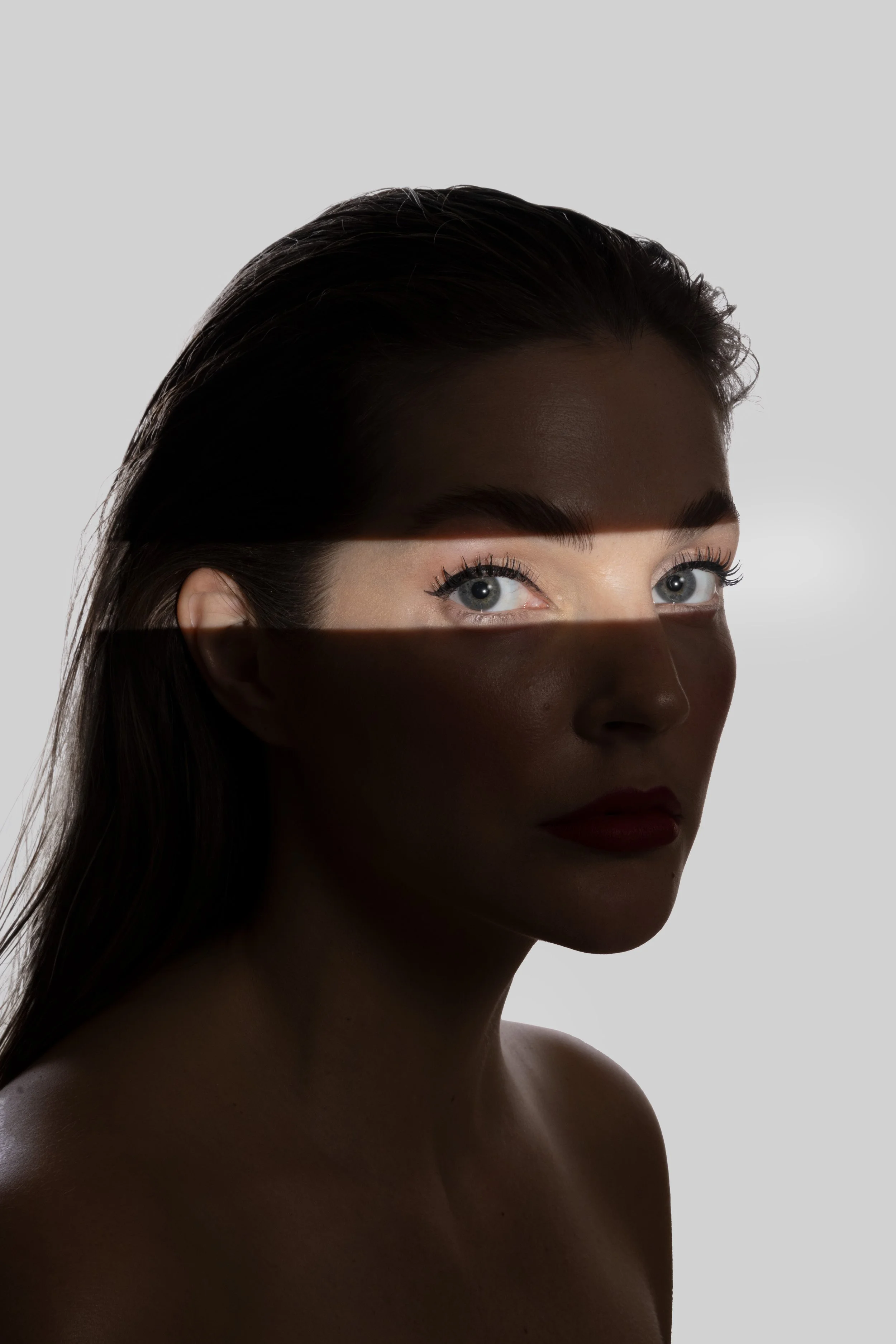 Ritratto frontale di una donna con pelle abbronzata, capelli castano scuro lisci e occhi chiari, con un'illuminazione contrastante che crea ombre sulla sua faccia.