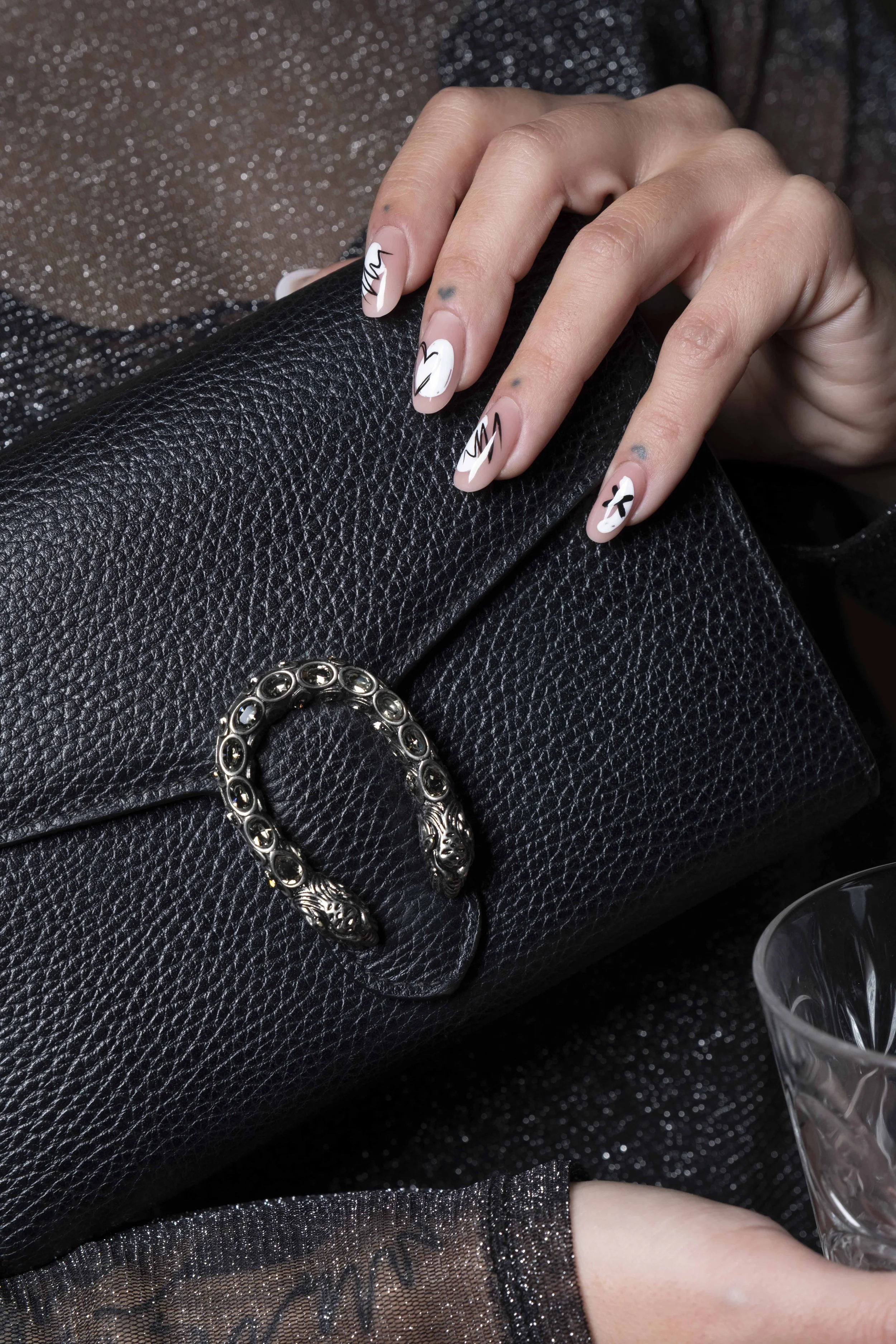 Manicure con design astratti su unghie di colore bianco, mani che riposano su una borsa nera in pelle con decorazione in metallo, sfondo con tessuto glitterato.