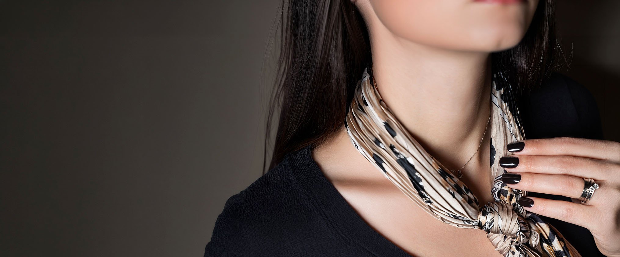 Primo piano di una donna con accessori eleganti, tra cui anelli, collana, smalto nero e foulard beige con motivi leopardati, mentre si sfiora il collo con la mano