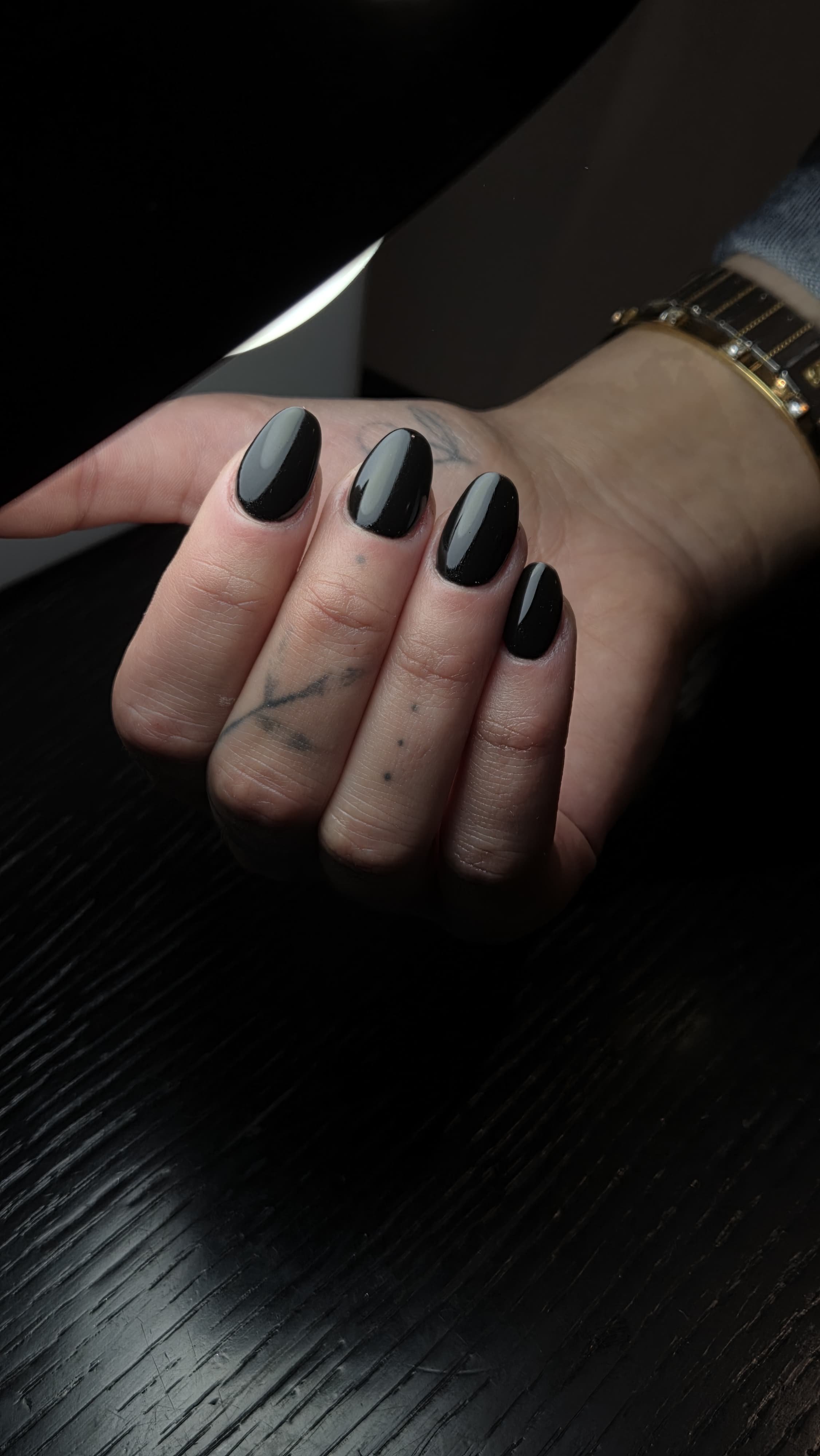 Unghia di mano dipinta di nero lucido, con tatuaggi visibili su dita e polso, indossando un orologio con cinturino in pelle.