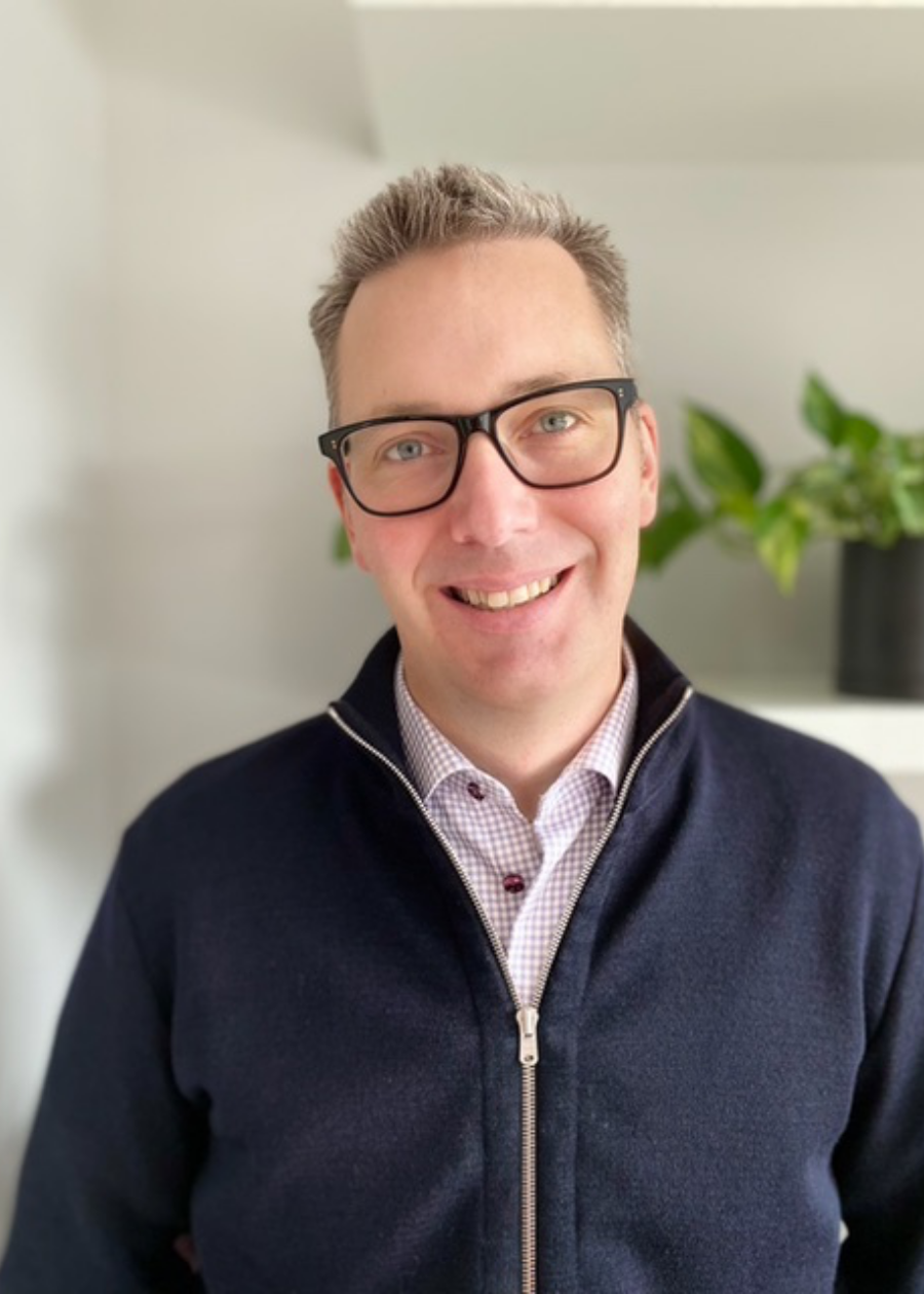 A smiling man wearing glasses, a checkered shirt, and a dark zip-up sweater in an indoor setting with a potted plant in the background.
