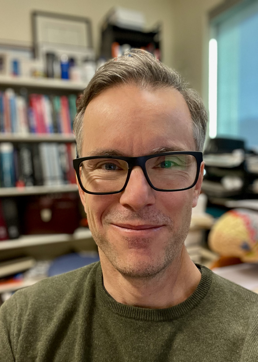 A man with short gray hair and glasses smiling at the camera indoors, with bookshelves and office supplies in the background.
