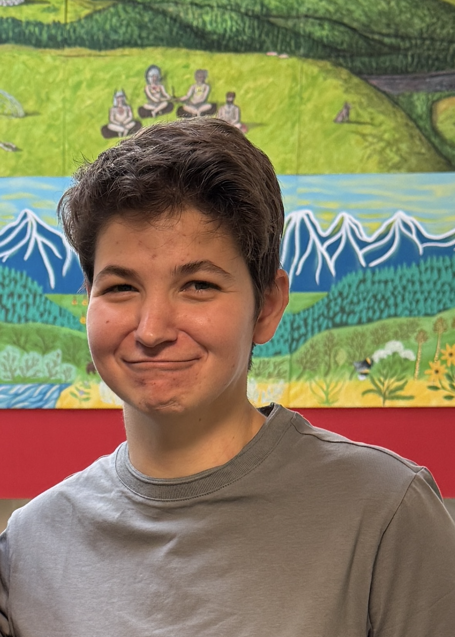 Young man with dark hair smiling in front of a colorful mural depicting mountains, fields, water, and people sitting cross-legged.