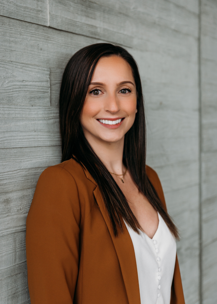 A woman with long dark hair and a brown blazer smiling while leaning against a wooden wall.