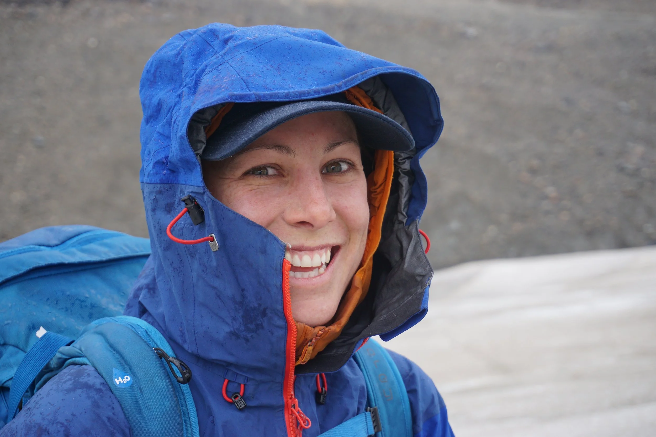 Close-up of a smiling person wearing a blue hooded jacket and cap, with a backpack, outdoors in a snowy or icy environment.