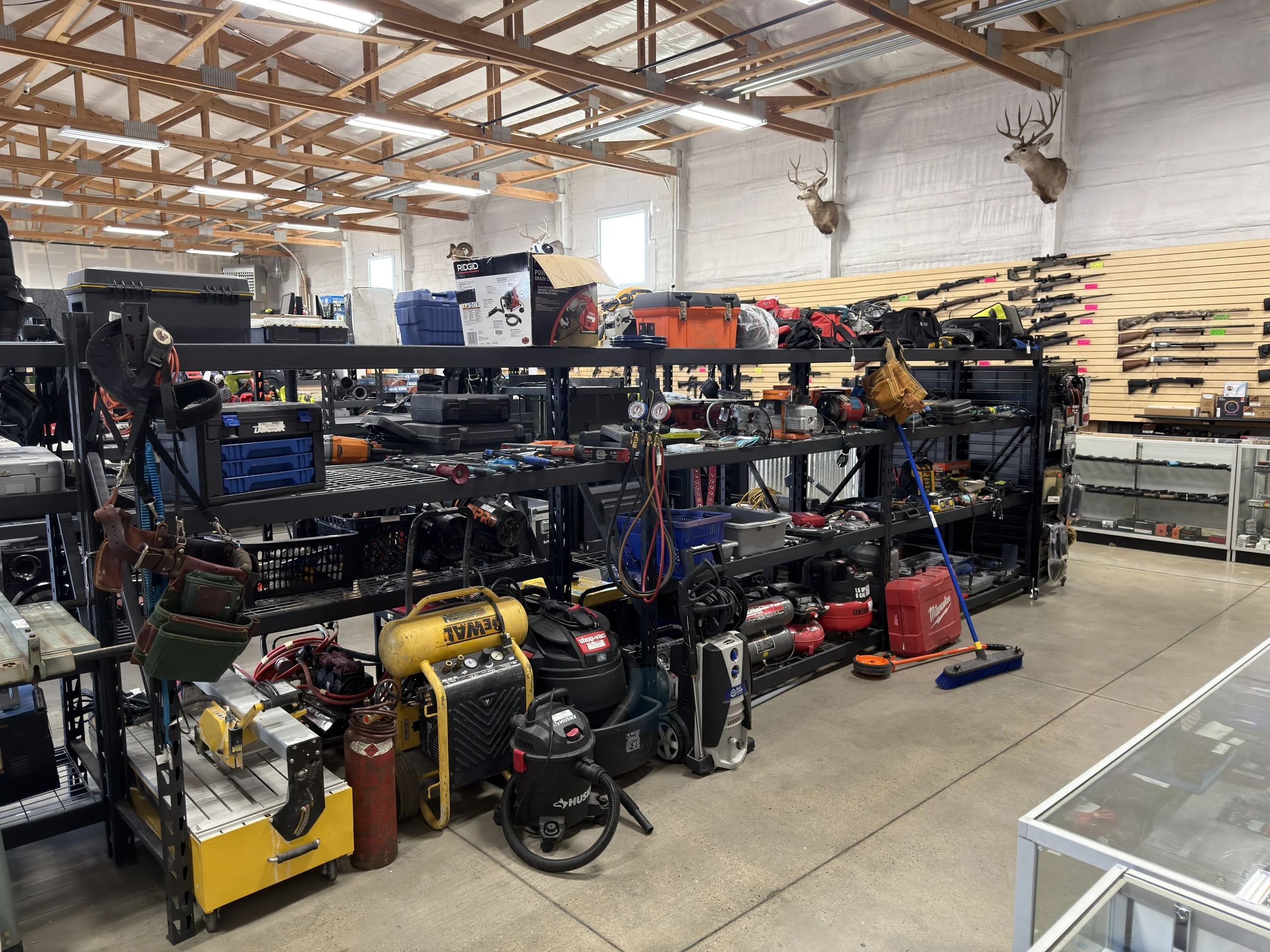 The image shows the inside of a hardware or tool store with shelves filled with power tools, equipment, and supplies. The top edge of the image reveals a ceiling with exposed wooden beams and fluorescent lighting. Mounted on the wall are several mounted deer heads with antlers, and there is a display of rifles on a wall along with pink and green price tags or labels. The foreground has various tools and equipment, including air compressors, and there is a broom leaning against the shelving unit.