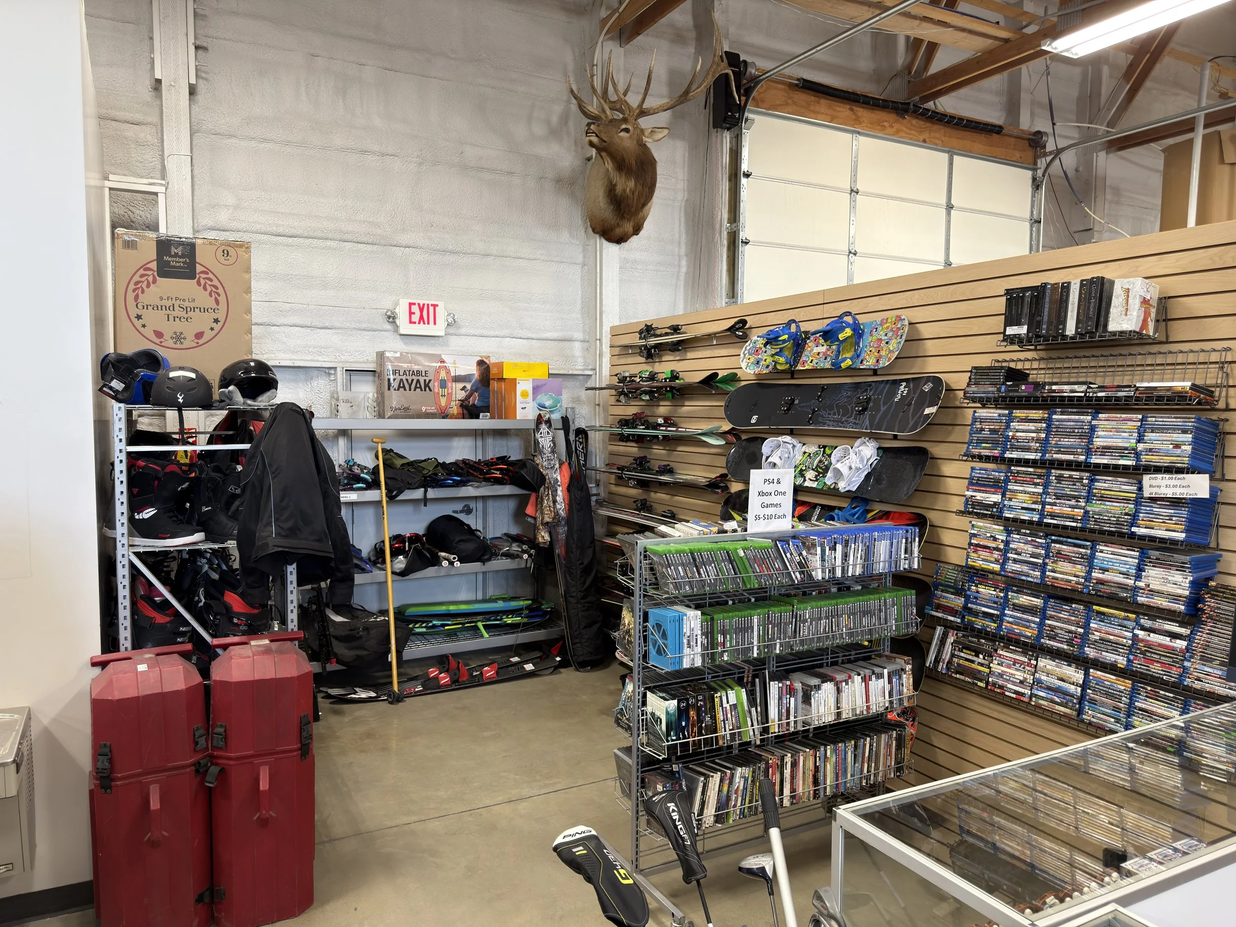 A retail store or pawn shop in Clearfield, UT interior with sports equipment, tools, jewelry, and decorative items. Prominently features a mounted deer head on the wall, various skateboards, video game discs, and stacked DVDs.