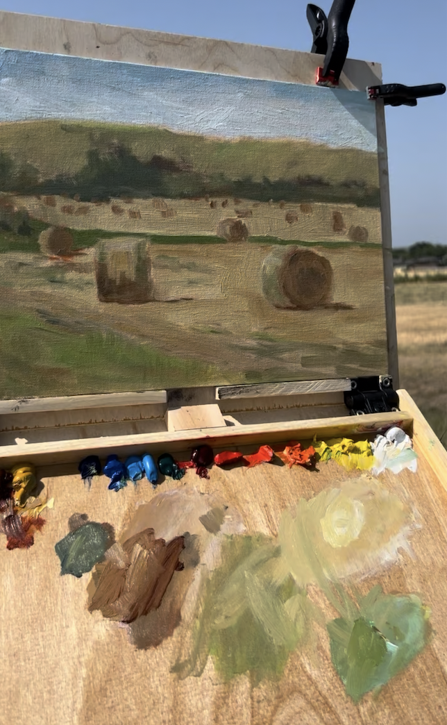Landscape painting depicting hay bales in a field with distant trees and a blue sky, on an easel outdoors.