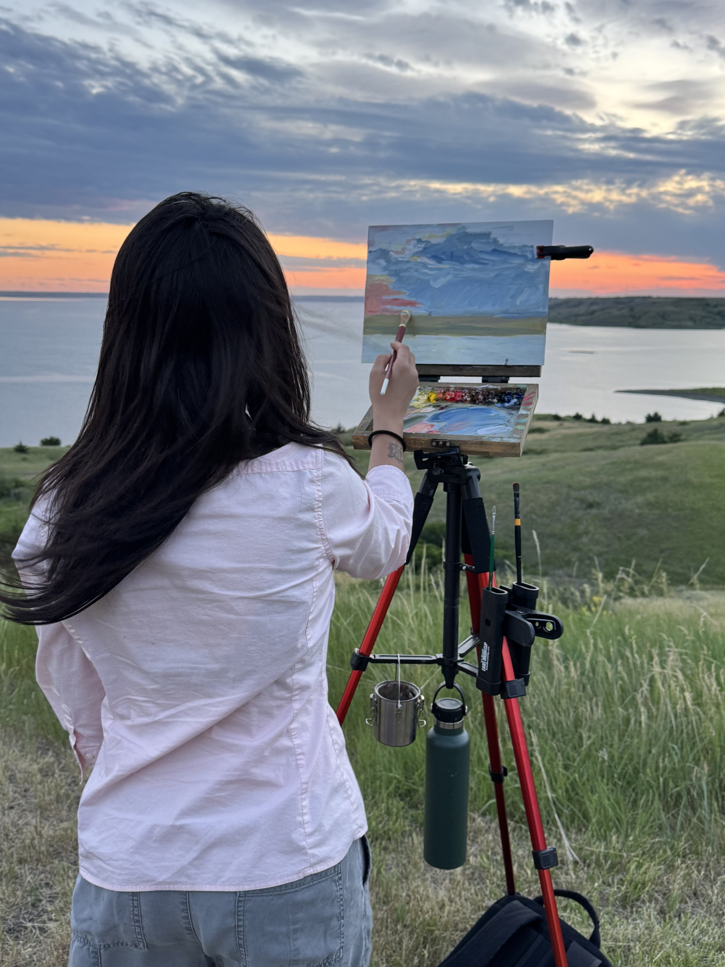 Person painting a landscape on a canvas using a tripod easel outdoors during sunset, with a body of water and a cloudy sky in the background.