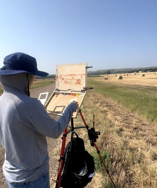 A person wearing a blue hat and gray hoodie painting a landscape scene on an easel outdoors in a field with hay bales under a clear blue sky.