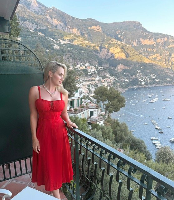 A woman in a red dress standing on a balcony overlooking a coastal town with boats on the water and mountains in the background.