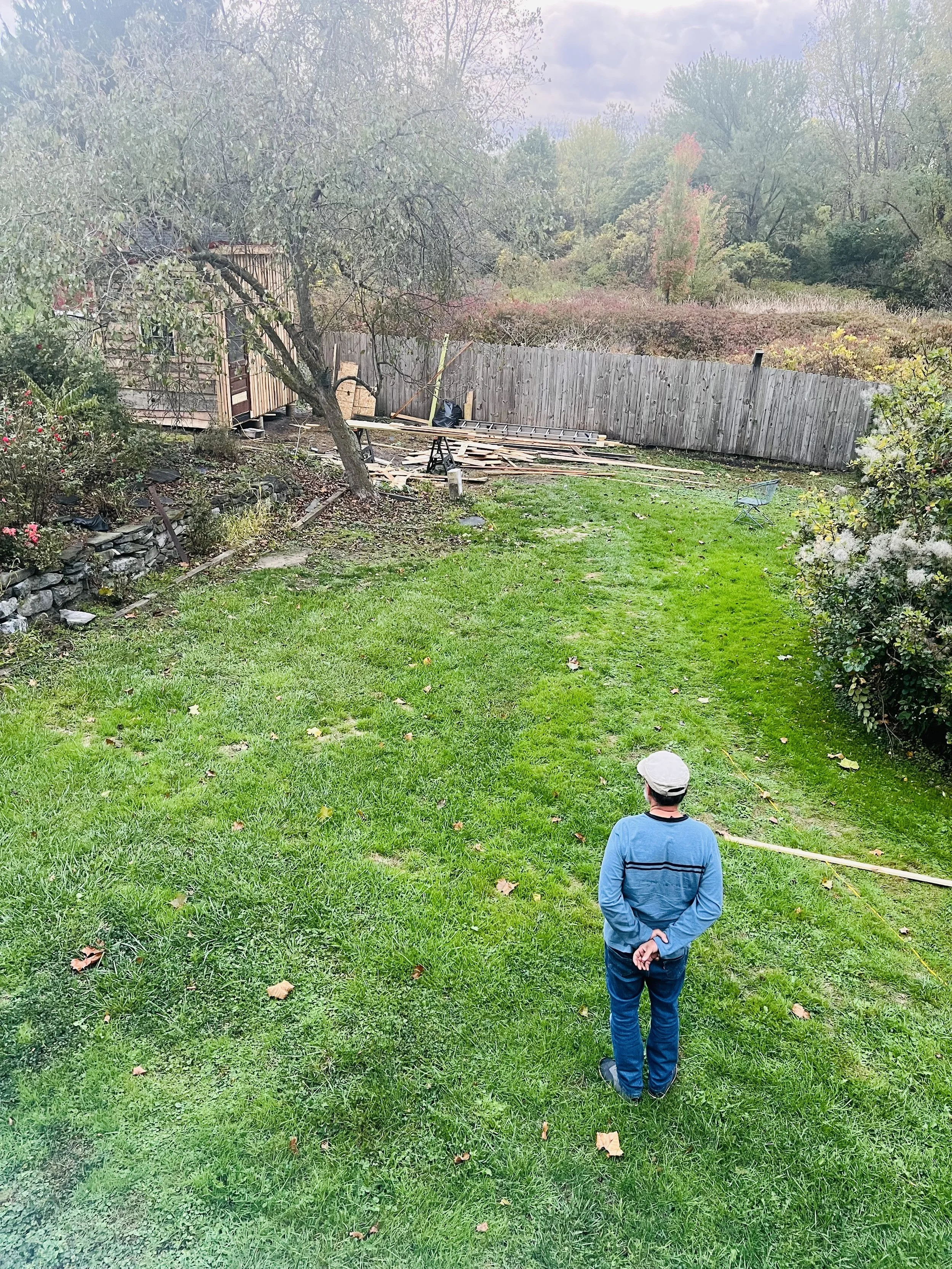 A person in a blue sweater and cap stands in a grassy backyard, facing away from the camera, with trees, bushes, and a wooden fence in the background.