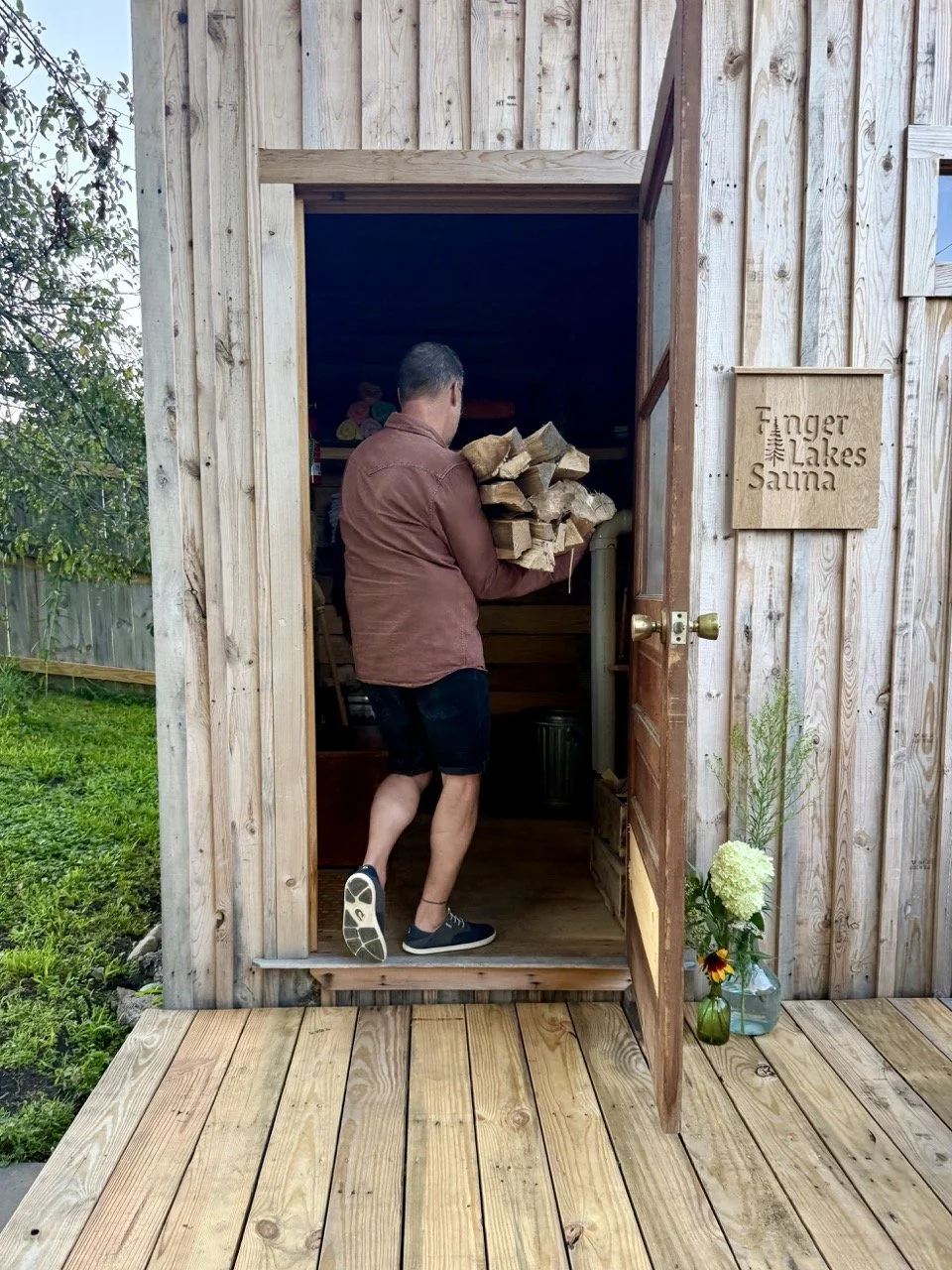 A man carrying firewood stands going through the entrance of a wooden sauna, with a sign that reads 'Finger Lakes Sauna' on the exterior wall. There are flowers in vases outside near the door, and the surrounding area is grassy with a wooden fence.